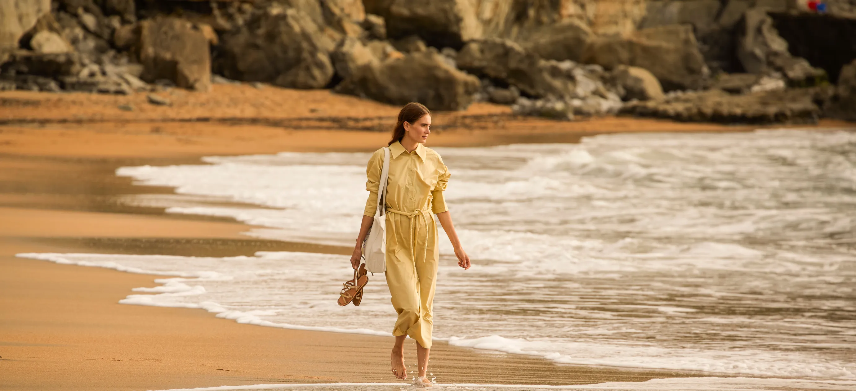 Donna in abito giallo che cammina a piedi nudi su una spiaggia sabbiosa con sandali e una borsa bianca.