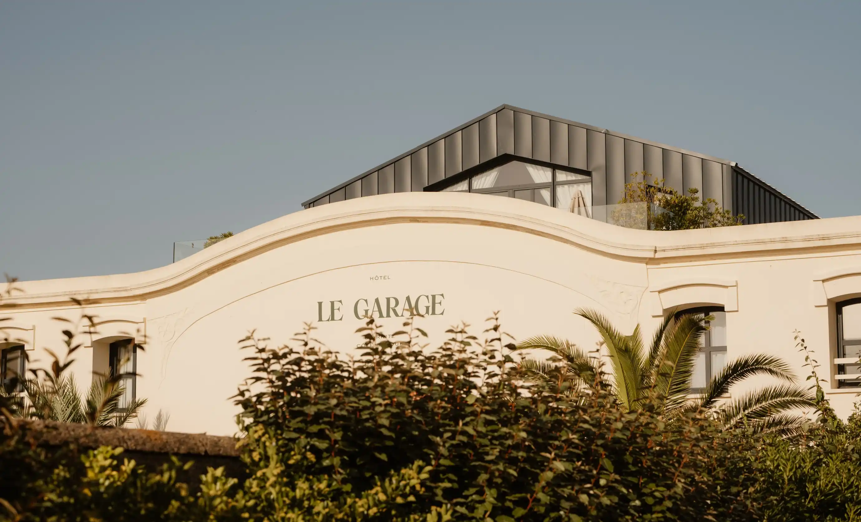 Hotel Le Garage con tetto moderno, facciata color crema e vegetazione di fronte sotto un cielo limpido.