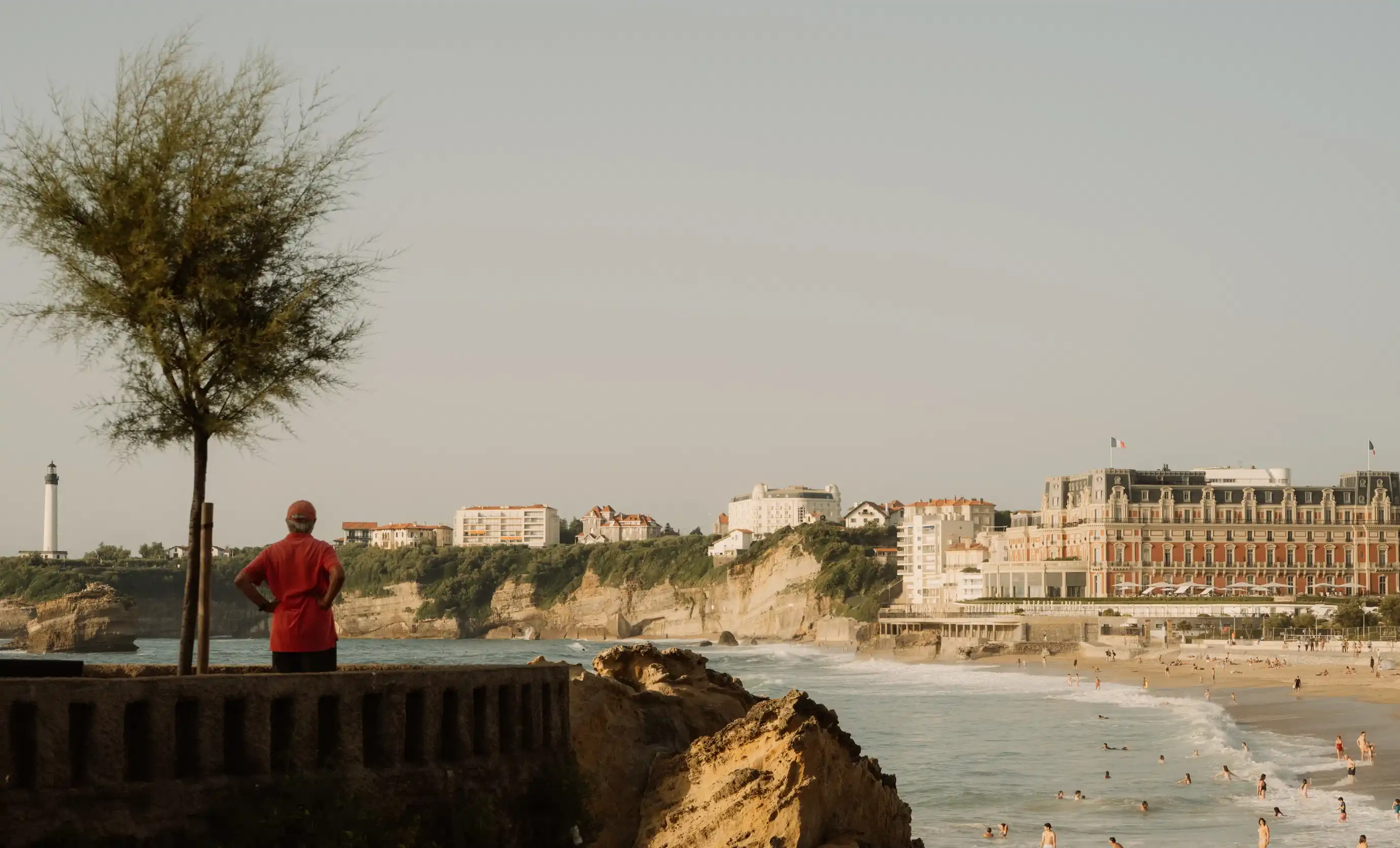 La persona con le mani sui fianchi si affaccia sulla spiaggia rocciosa con onde ed edifici sulle scogliere sotto un cielo limpido.