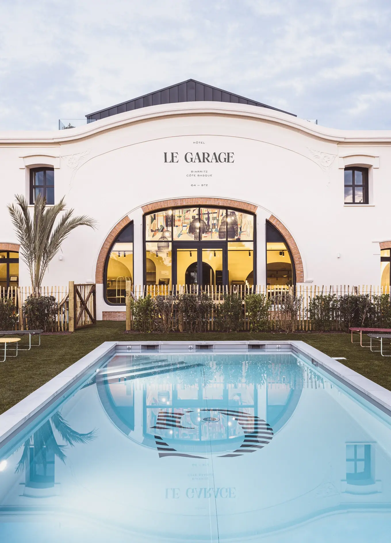 Edificio dell'Hotel Le Garage con grandi finestre ad arco che si riflettono in una chiara piscina rettangolare.