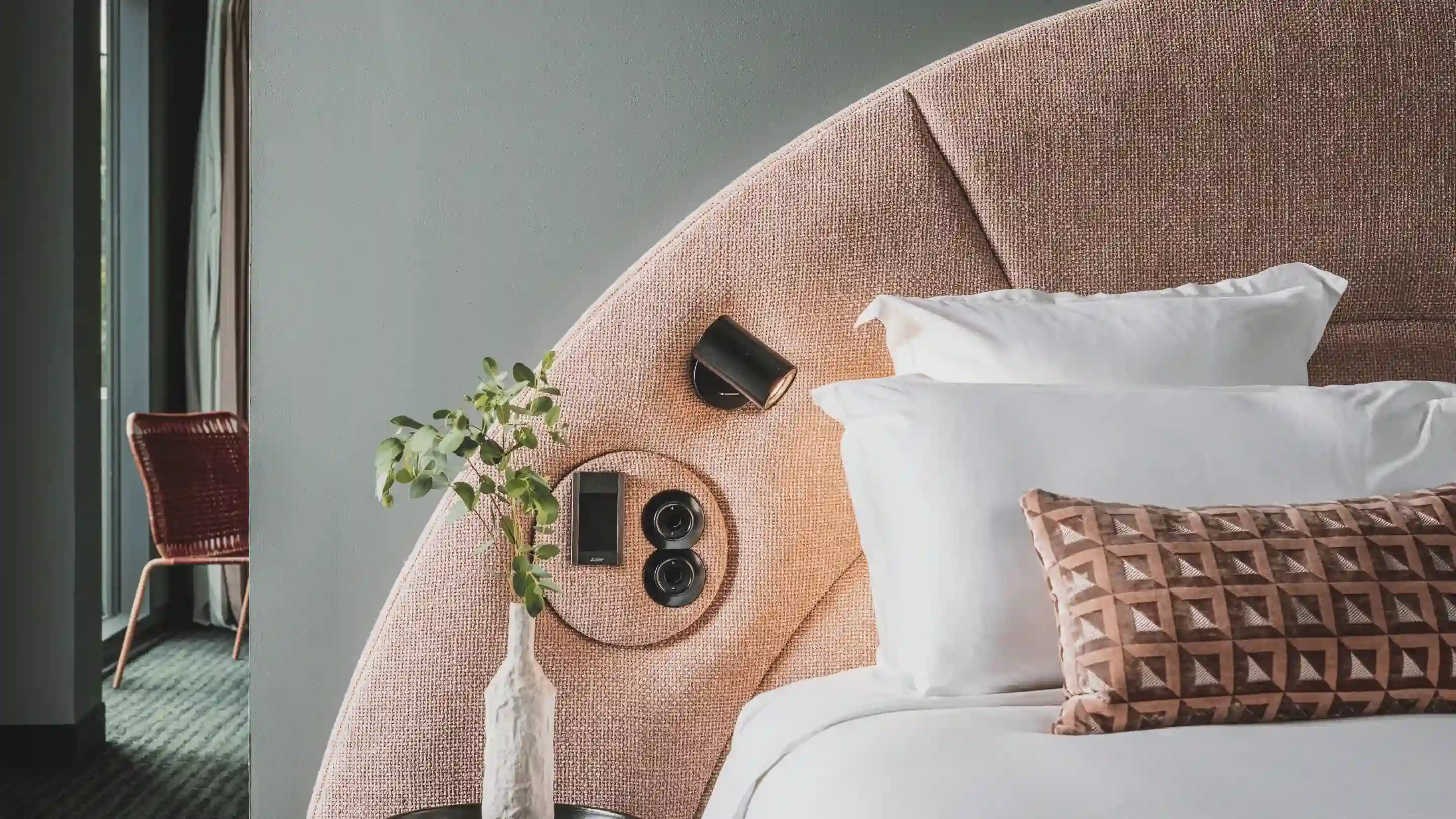 Letto moderno con testiera in tessuto curvato, cuscini bianchi, cuscino a motivi geometrici e pianta da comodino in vaso.