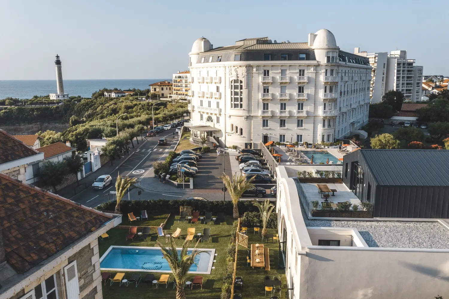 Hotel costiero con facciata bianca, piscine all'aperto, giardini, auto parcheggiate e un lontano faro in riva al mare.