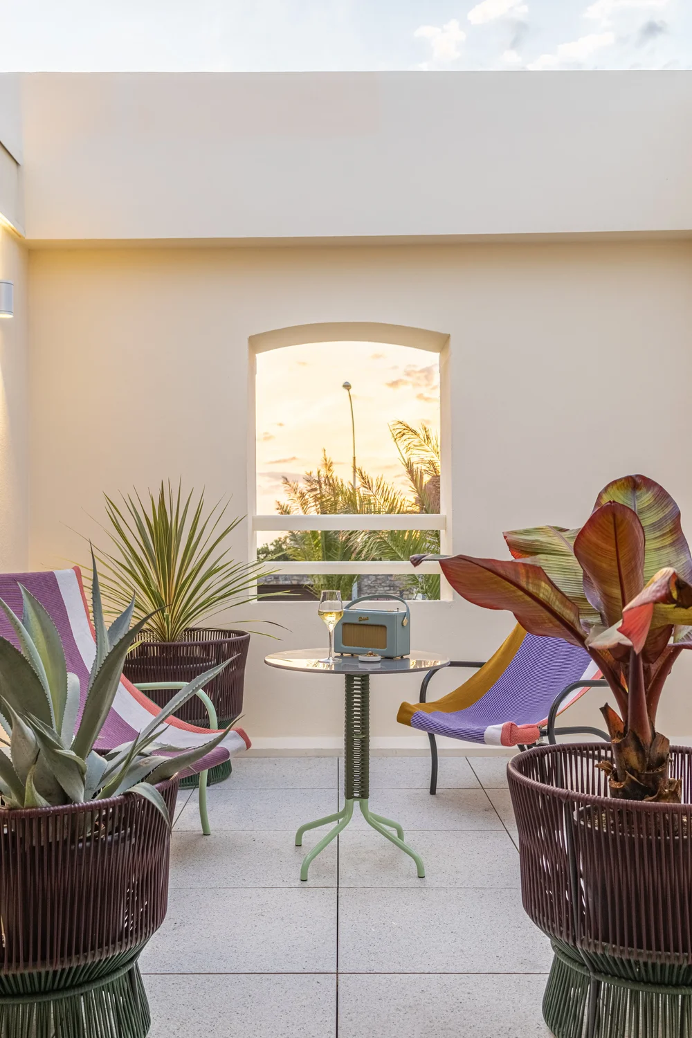 Terrazza minimalista con due sedie colorate, un tavolino, piante e un cielo al tramonto attraverso una finestra ad arco.