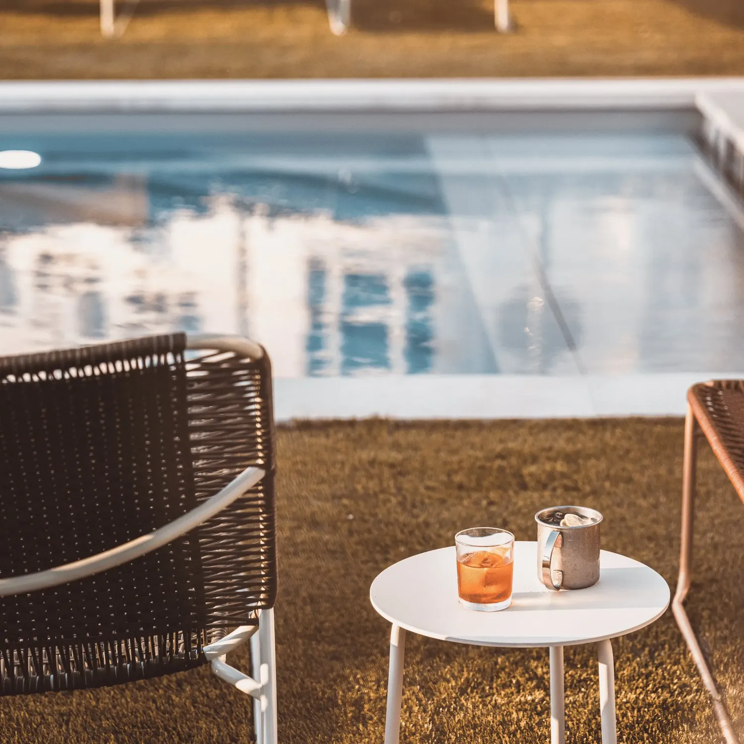Due drink su un tavolo rotondo bianco accanto a una piscina, con due sedie intrecciate sull'erba.