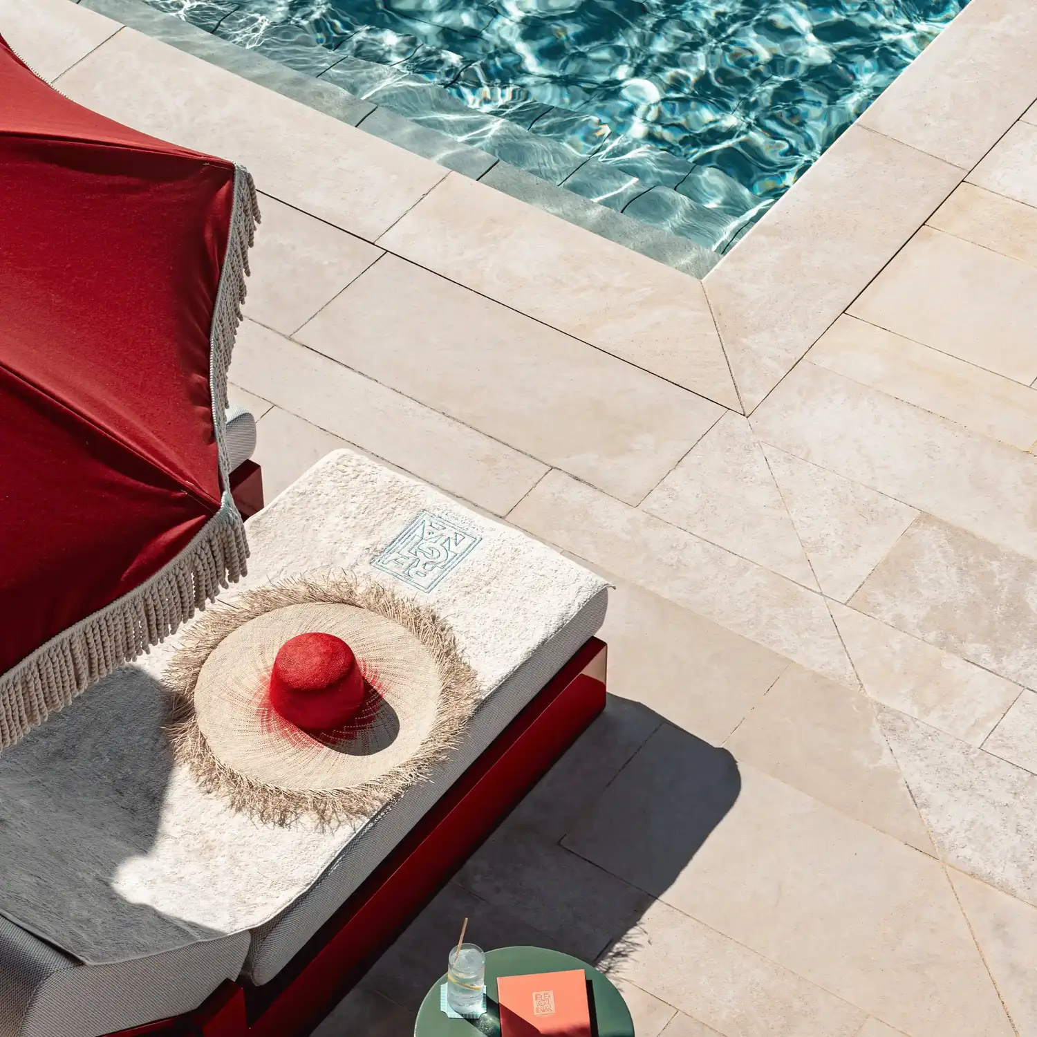 Poltrona a bordo piscina con un cappello da sole rosso e paglia sotto un ombrellone rosso e un tavolo con bibita e libro.