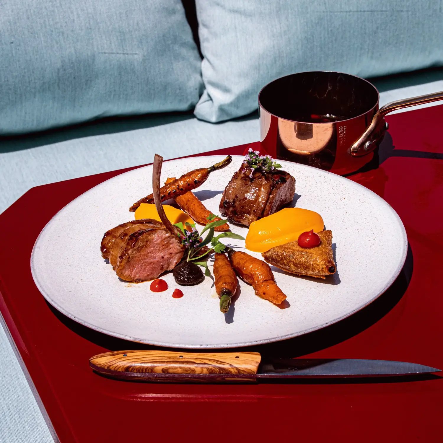 Impiattare con costolette di agnello cotte, carote grigliate, purea e guarnire su un tavolo rosso con coltello e pentola.