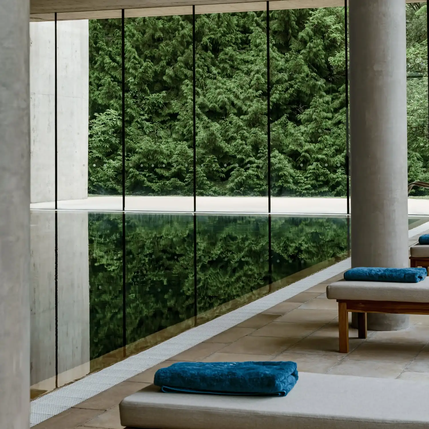 Piscina coberta com parede de vidro refletindo árvores verdes, piso de pedra e espreguiçadeiras com toalhas azuis.