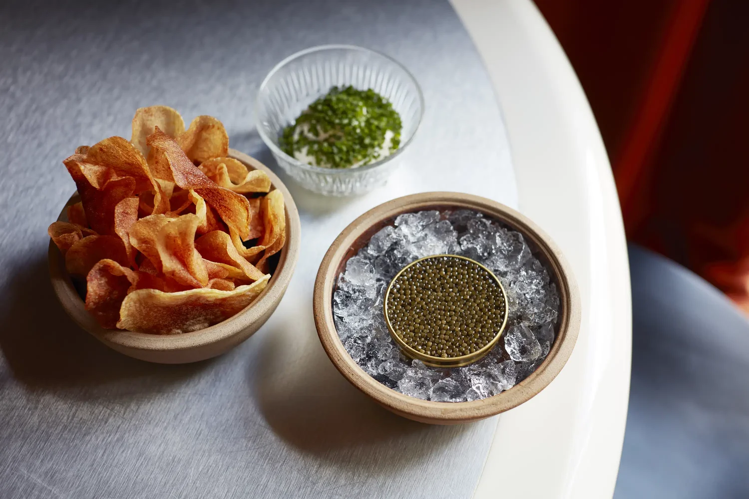 Tigela de batatas fritas, uma lata de caviar com gelo e um prato de creme com ervas picadas em uma mesa.
