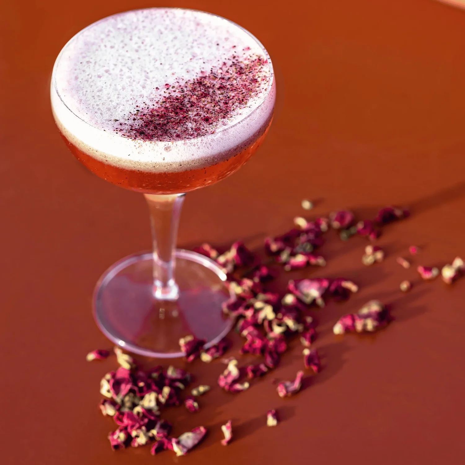 Coquetel com cobertura espumosa e granulado vermelho em um copo cupê, com pétalas de flores secas em uma superfície marrom.