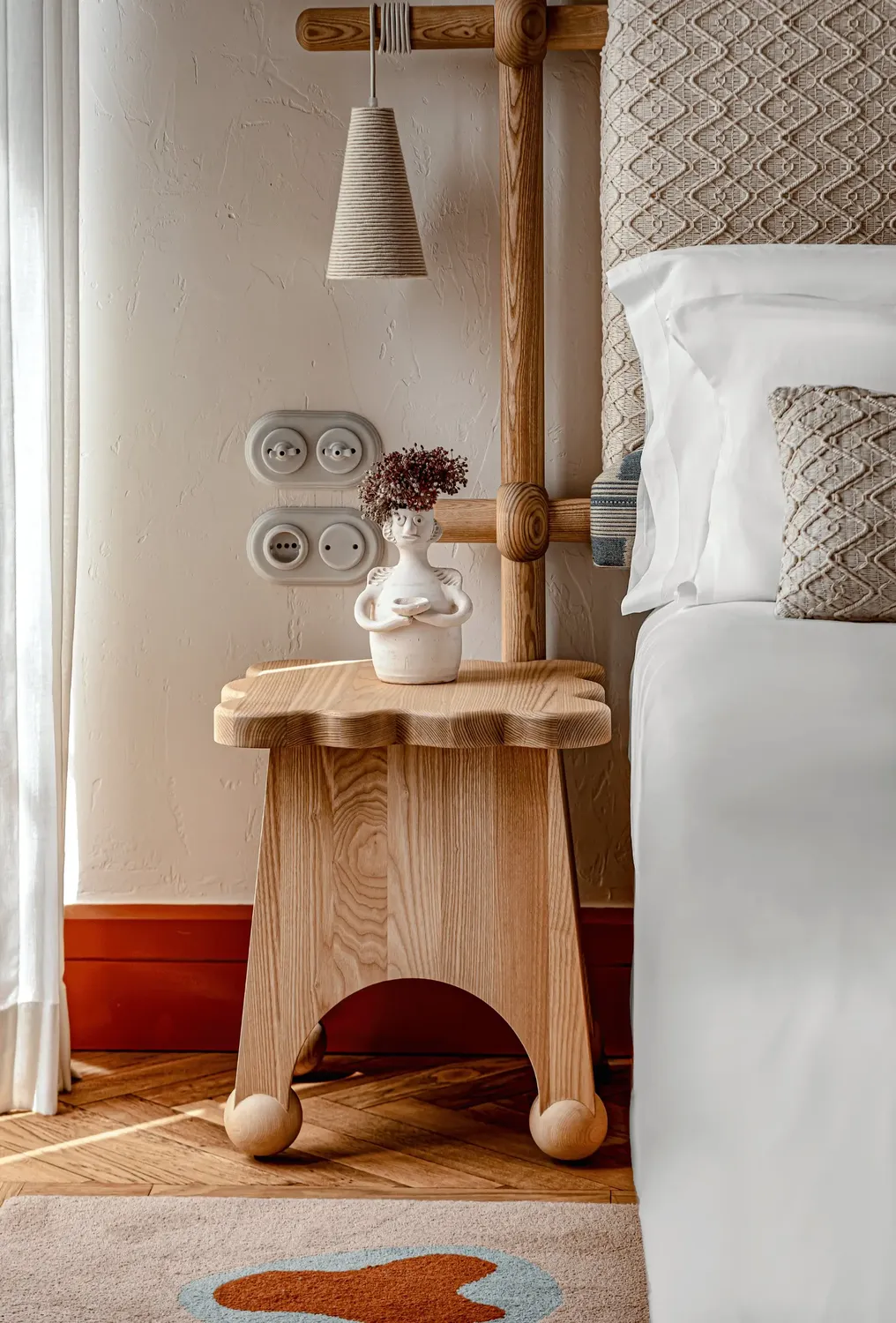 Mesa lateral de madeira com um vaso com flores secas ao lado de uma cama com almofadas texturizadas.