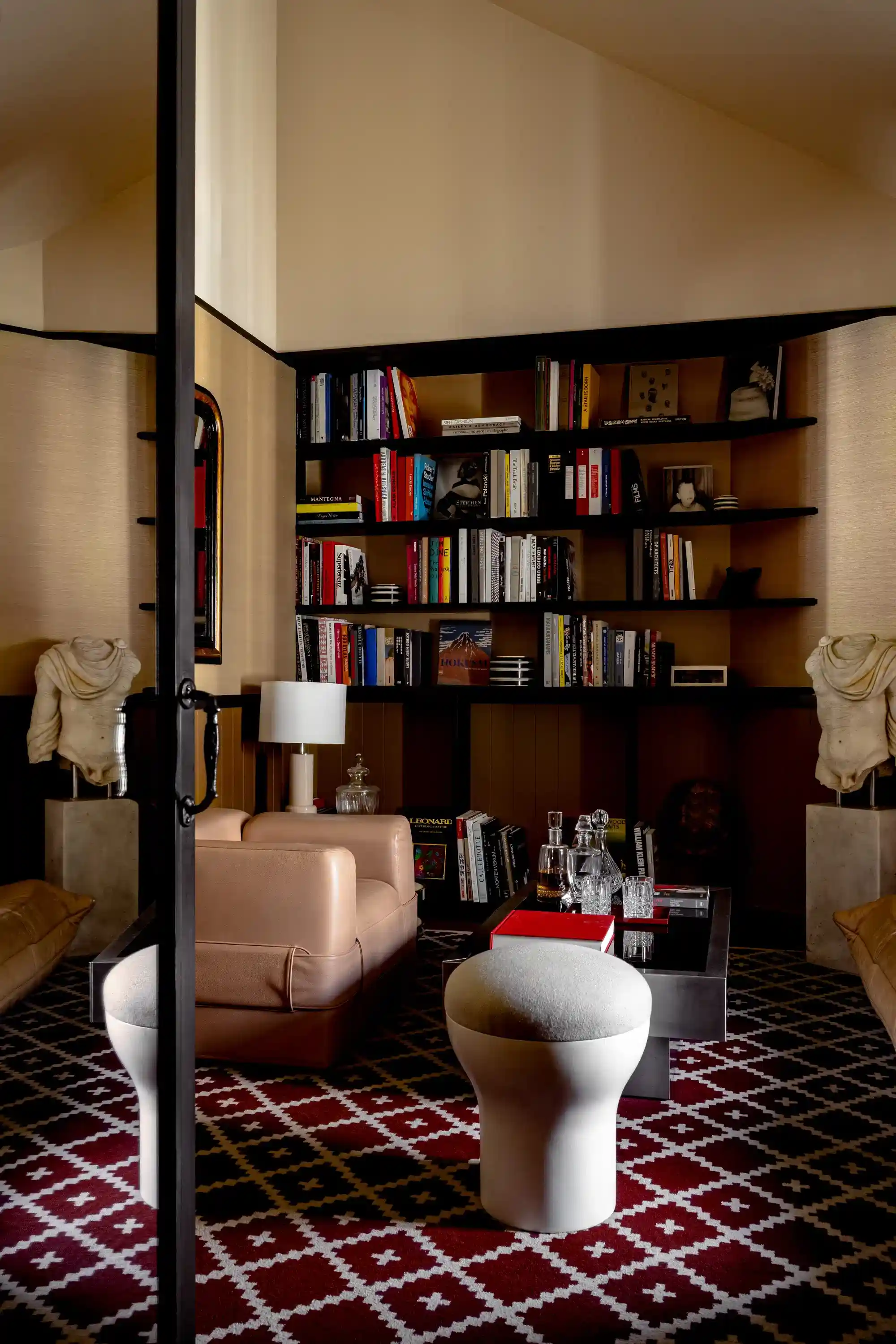 Accogliente sala biblioteca con poltrona in pelle, moquette a motivi geometrici, sgabello moderno, librerie e busti classici.