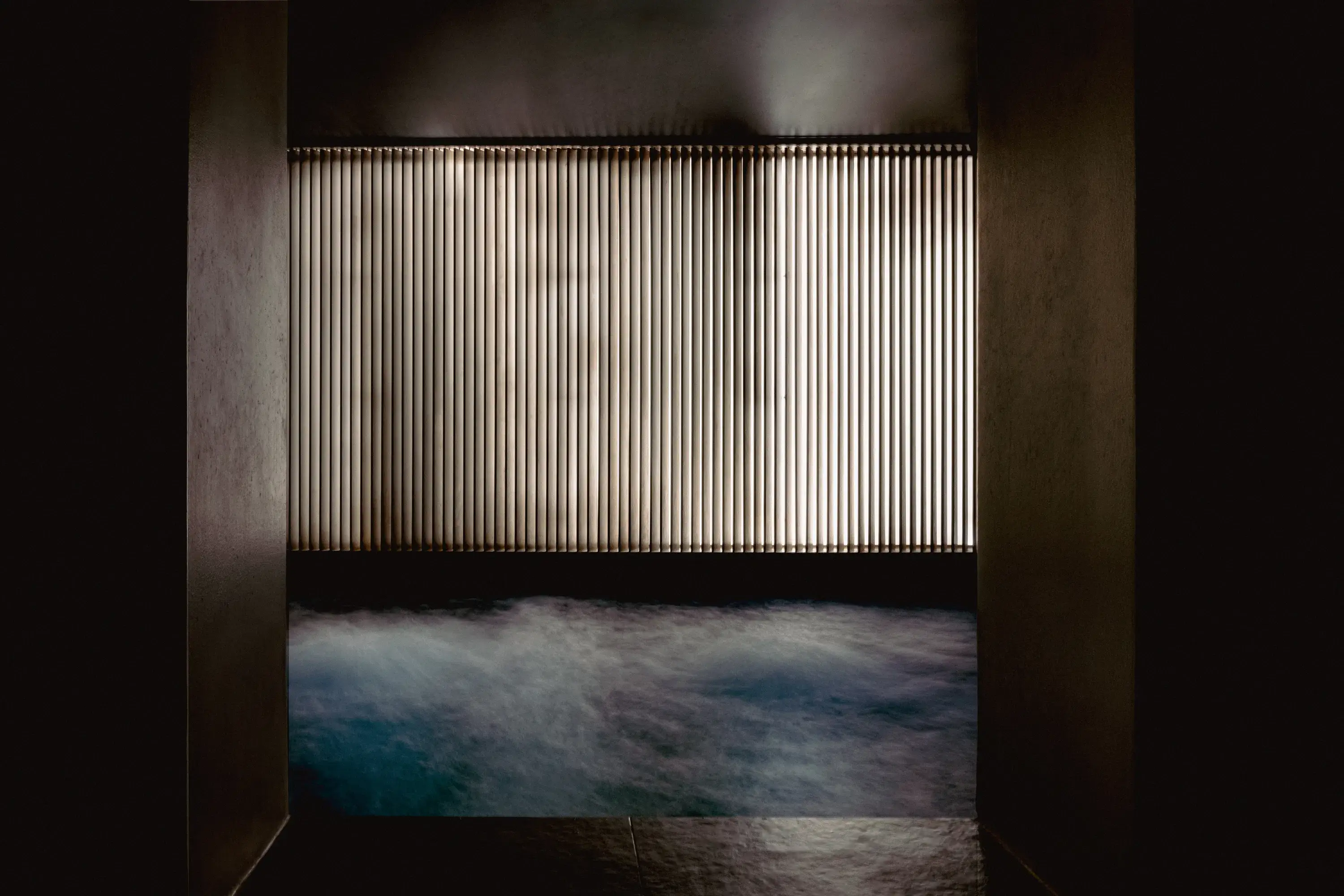 Piscina termale coperta poco illuminata con acqua gorgogliante davanti a un'alta parete a doghe verticali.