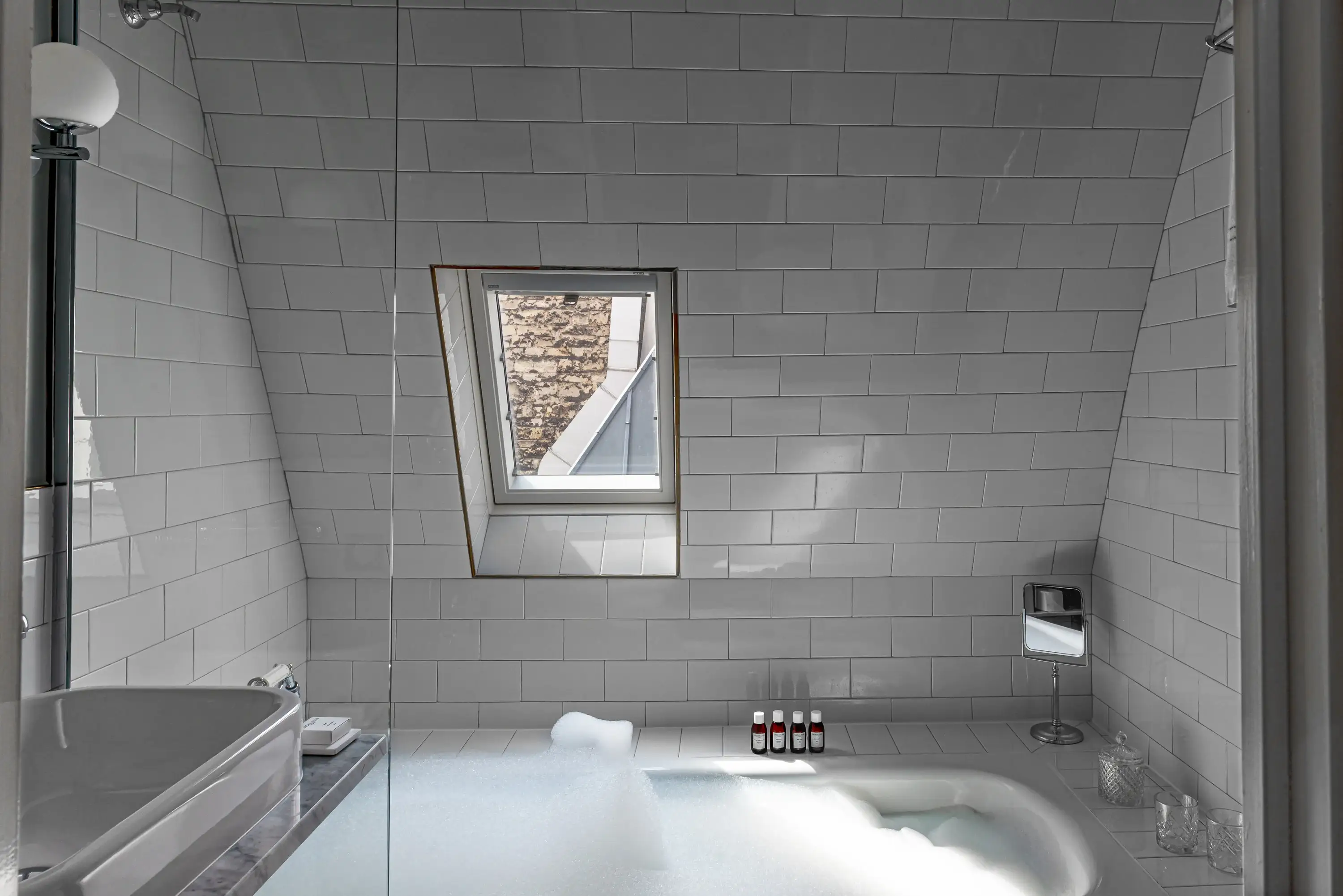 Banheiro moderno com banheira de espuma, janela pequena, azulejos brancos e produtos de higiene pessoal perto da banheira.