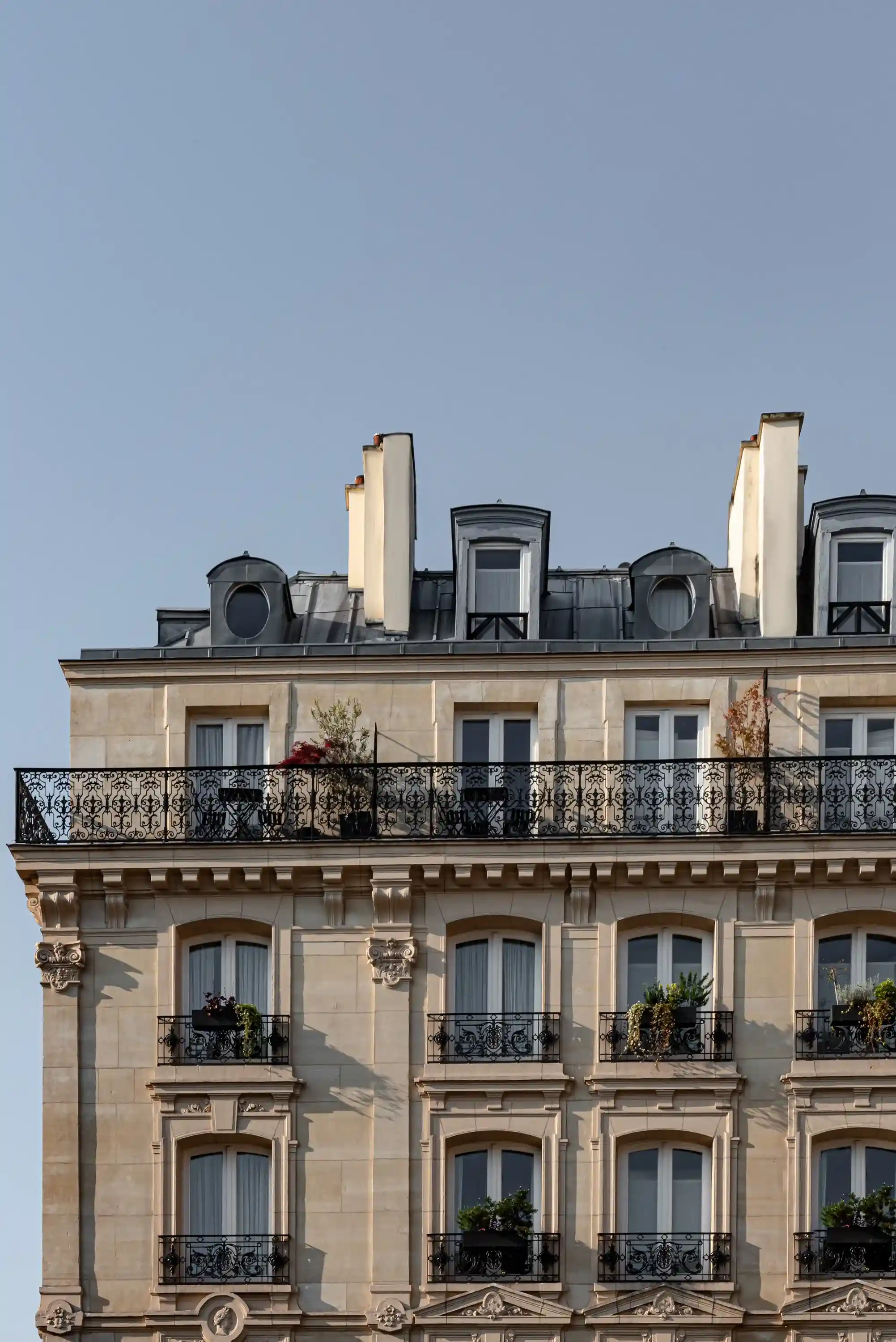 Fachada superior do edifício em estilo parisiense com varandas de ferro forjado e plantas sob um céu azul claro.