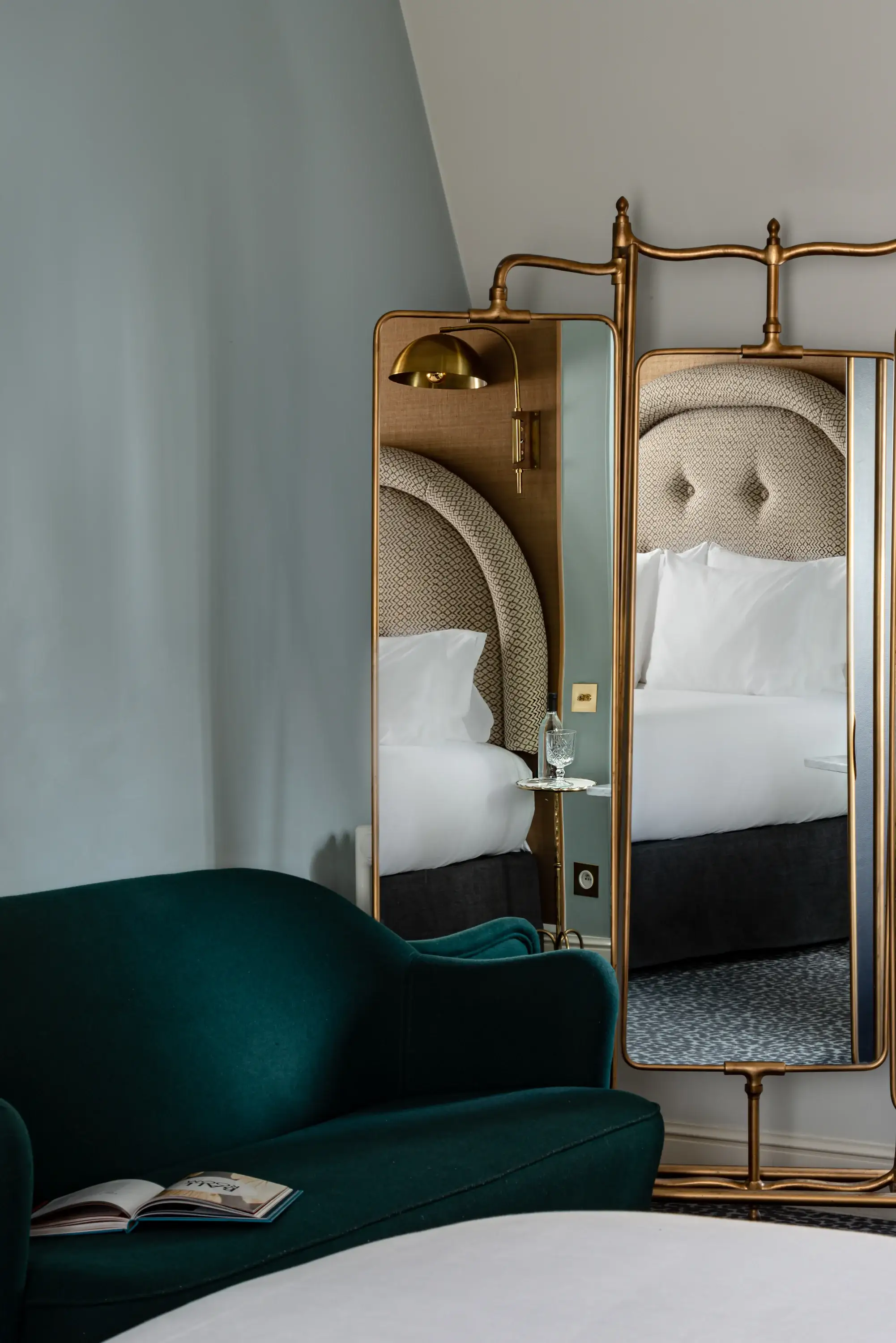 Sofá azul-petróleo com livro aberto em frente a um espelho de três painéis refletindo uma cama com cabeceira estampada.