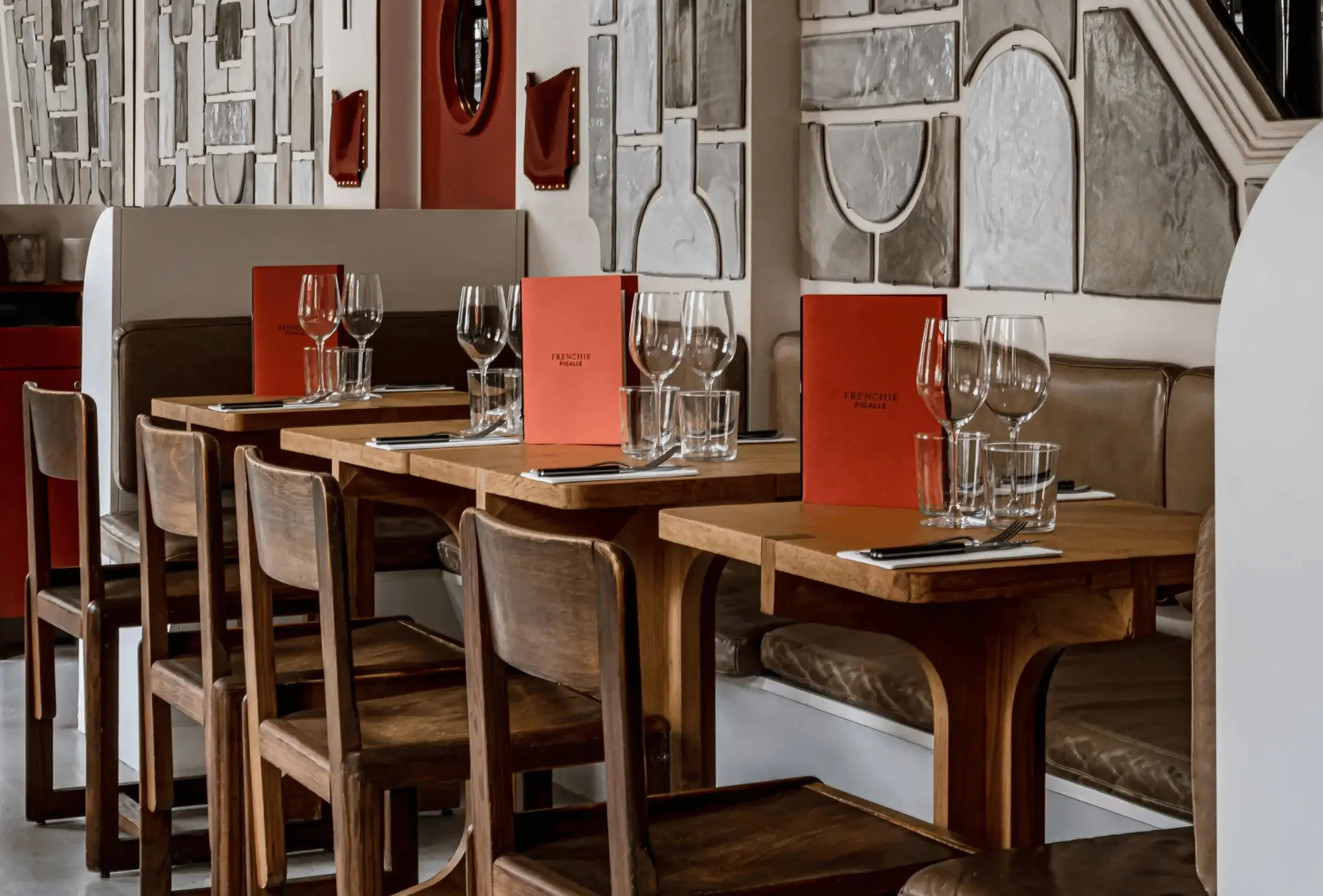 Mesas e cadeiras de madeira em um restaurante com menus, taças de vinho e arte de parede ao fundo.