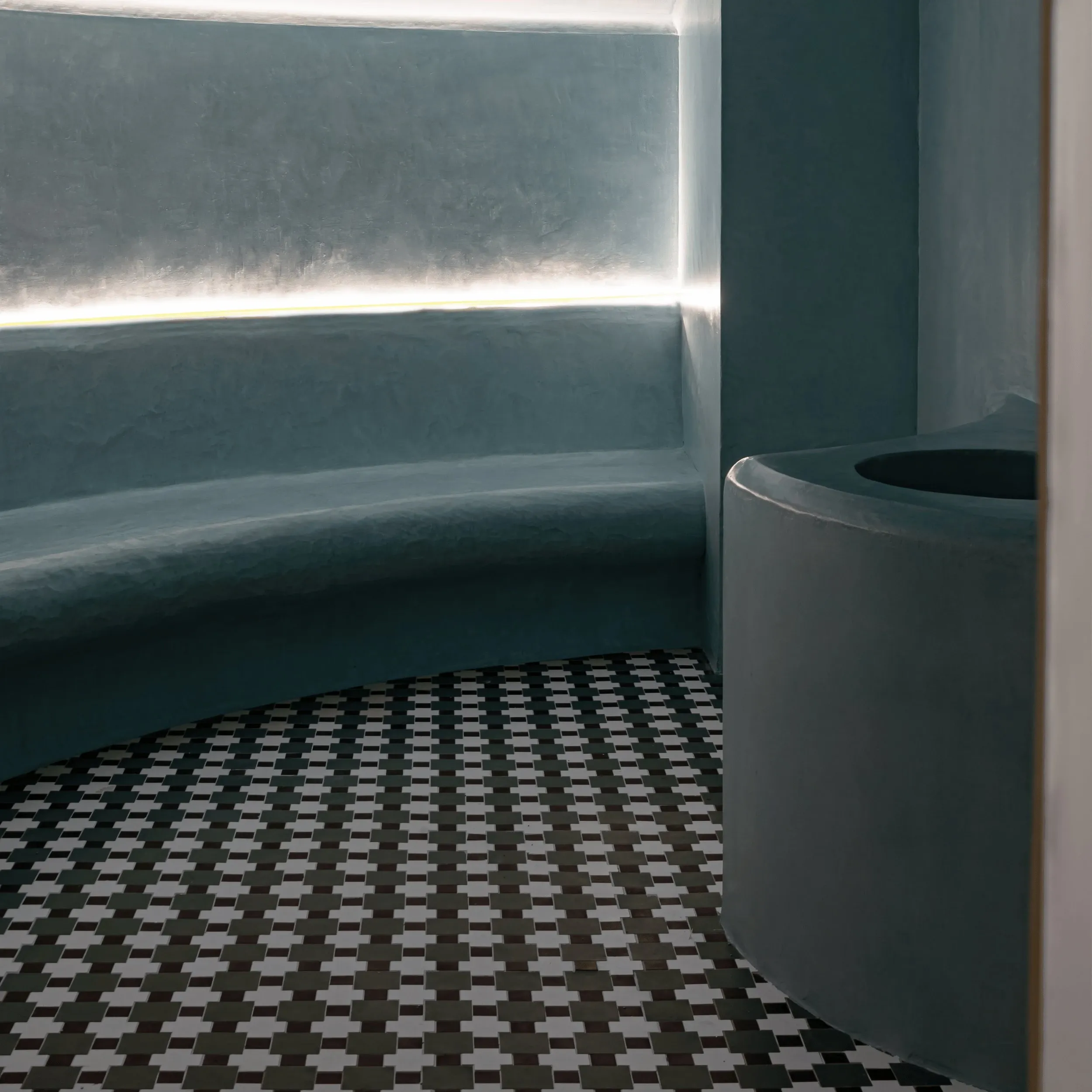 Panca e parete grigie curve con illuminazione a strisce sopra il pavimento piastrellato bianco e nero a motivi geometrici.