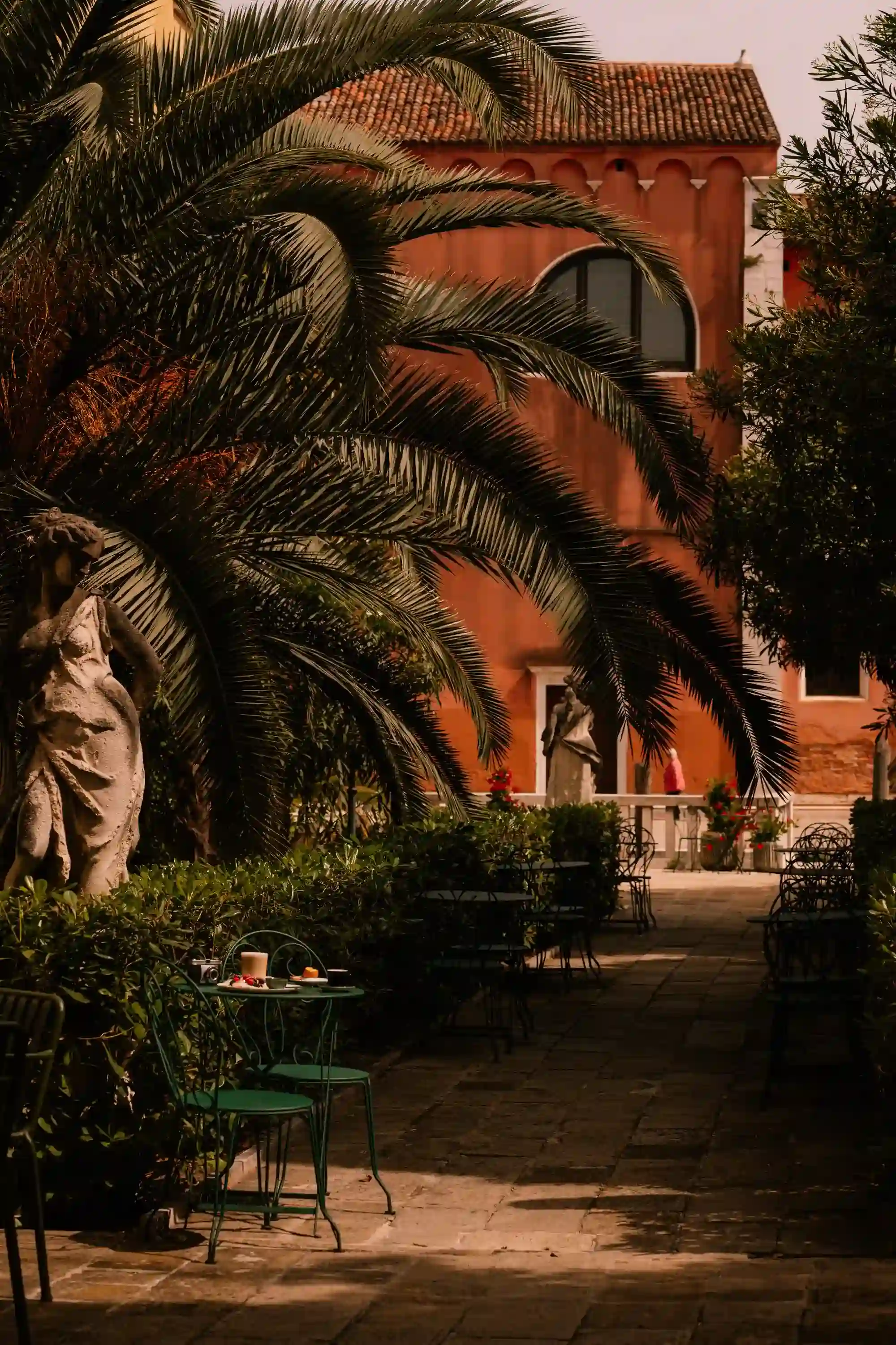 Mesas e cadeiras de café ao ar livre ao longo de um caminho sombreado com palmeiras e estátuas ao lado de um prédio de tijolos.