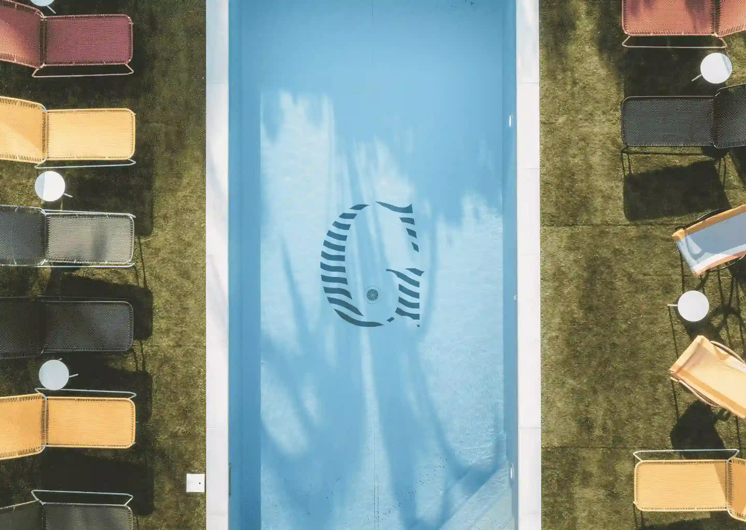 Veduta aerea di una piscina rettangolare con una grande lettera G nell'acqua, circondata da sedie a sdraio.
