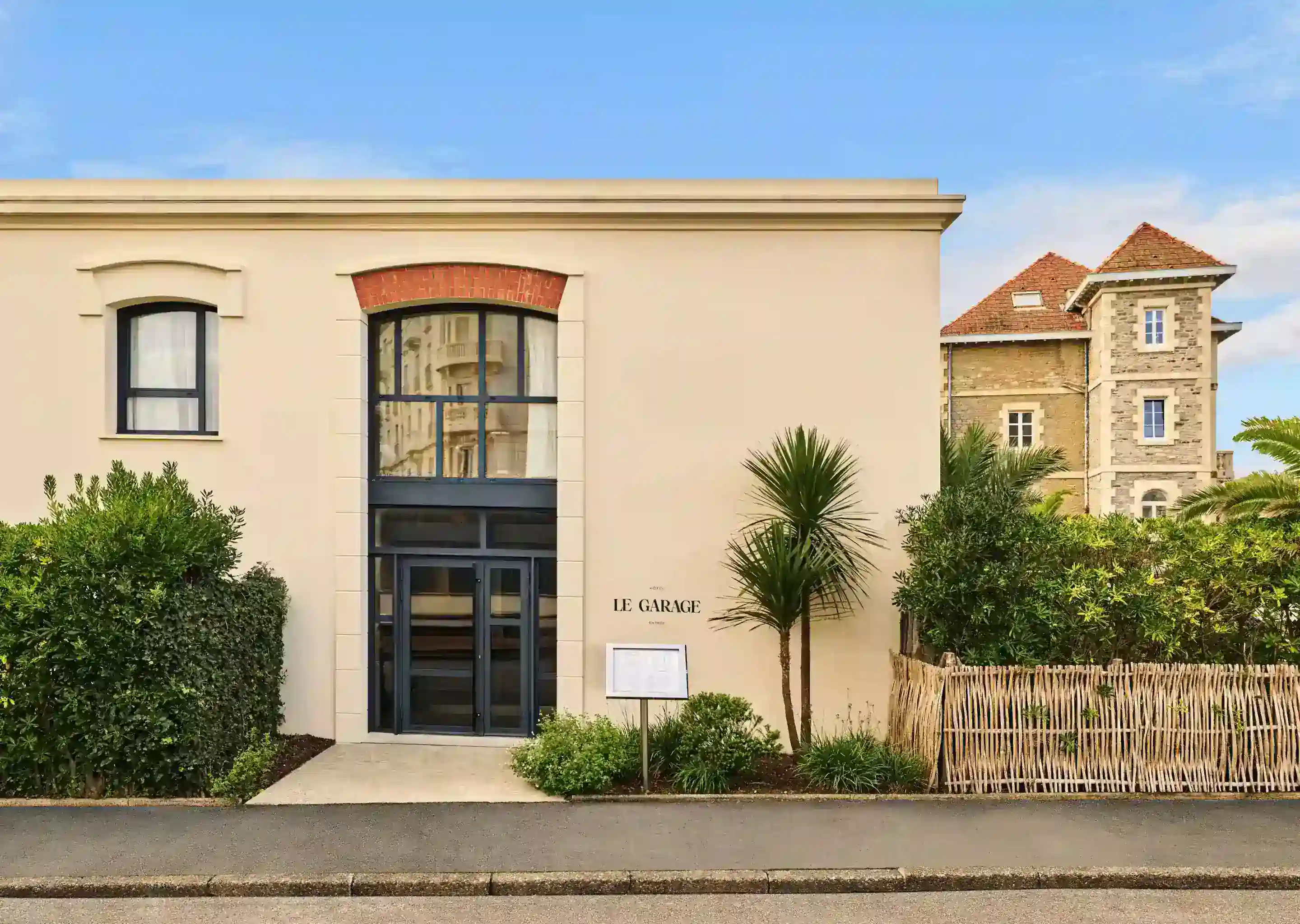 Edificio color crema con porte in vetro con cornice nera, cartello con la scritta Le Garage e vegetazione intorno all'ingresso.