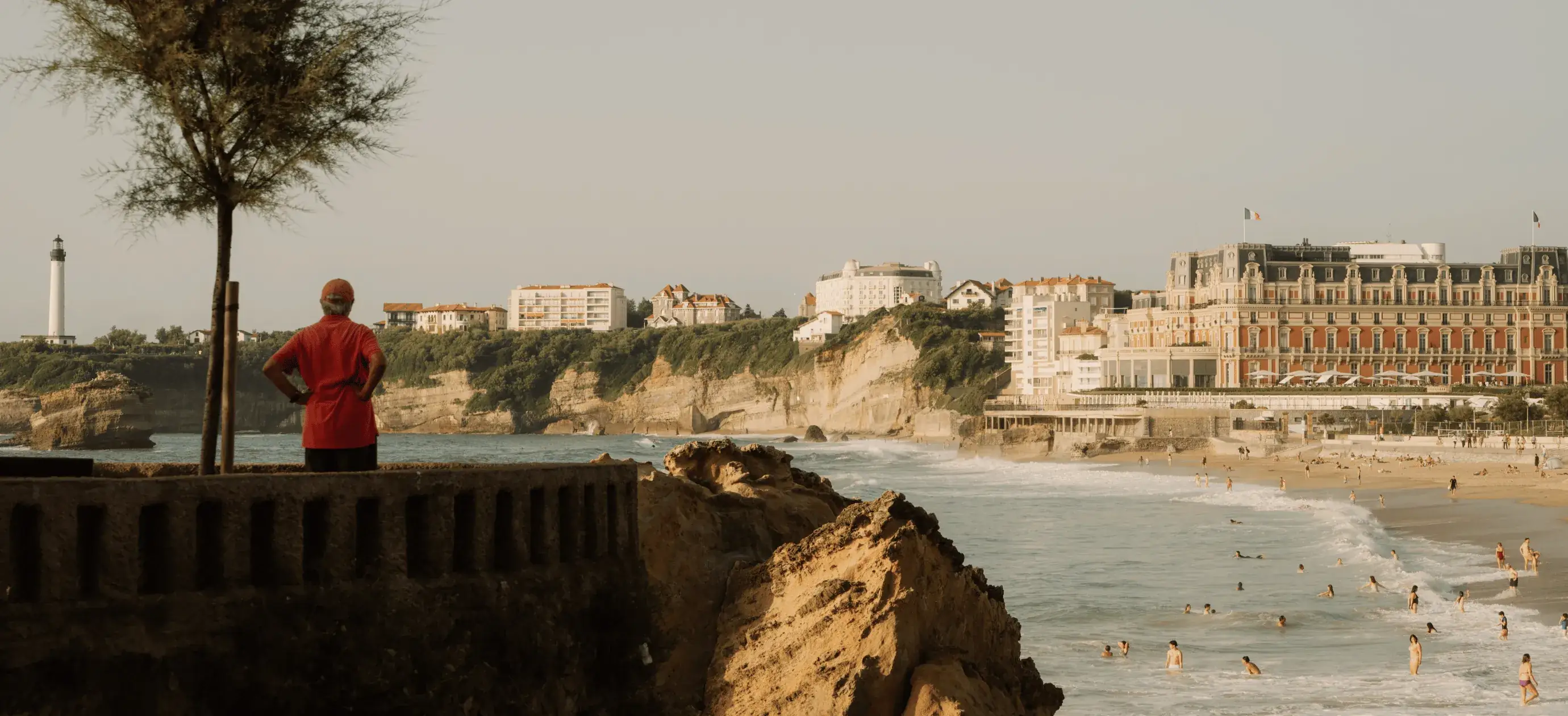 Persona in camicia rossa in piedi vicino a un albero con vista sulla spiaggia, sull'oceano, sulle scogliere e sul grande hotel storico.