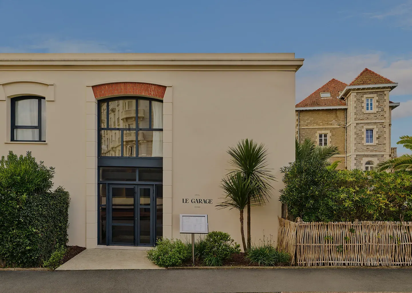 Esterno dell'Hotel Le Garage con porta a vetri, arbusti, palme e vecchio edificio in pietra sullo sfondo.