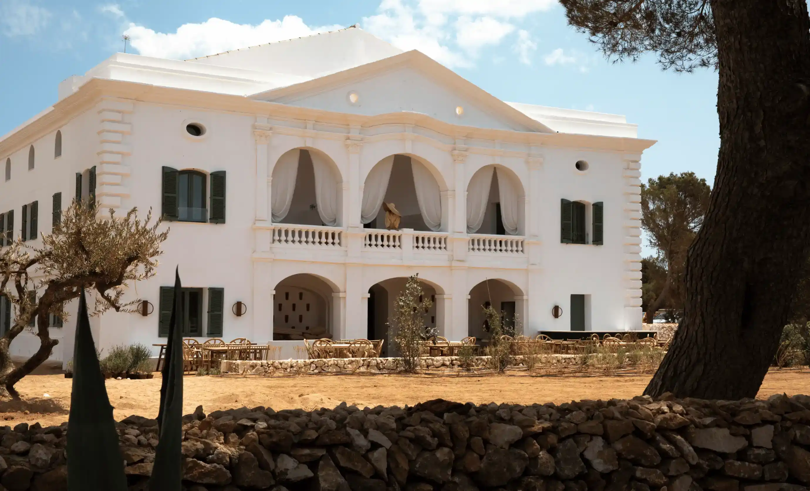 Edificio mediterraneo bianco con balconi ad arco, posti a sedere all'aperto e alberi davanti sotto il cielo blu.