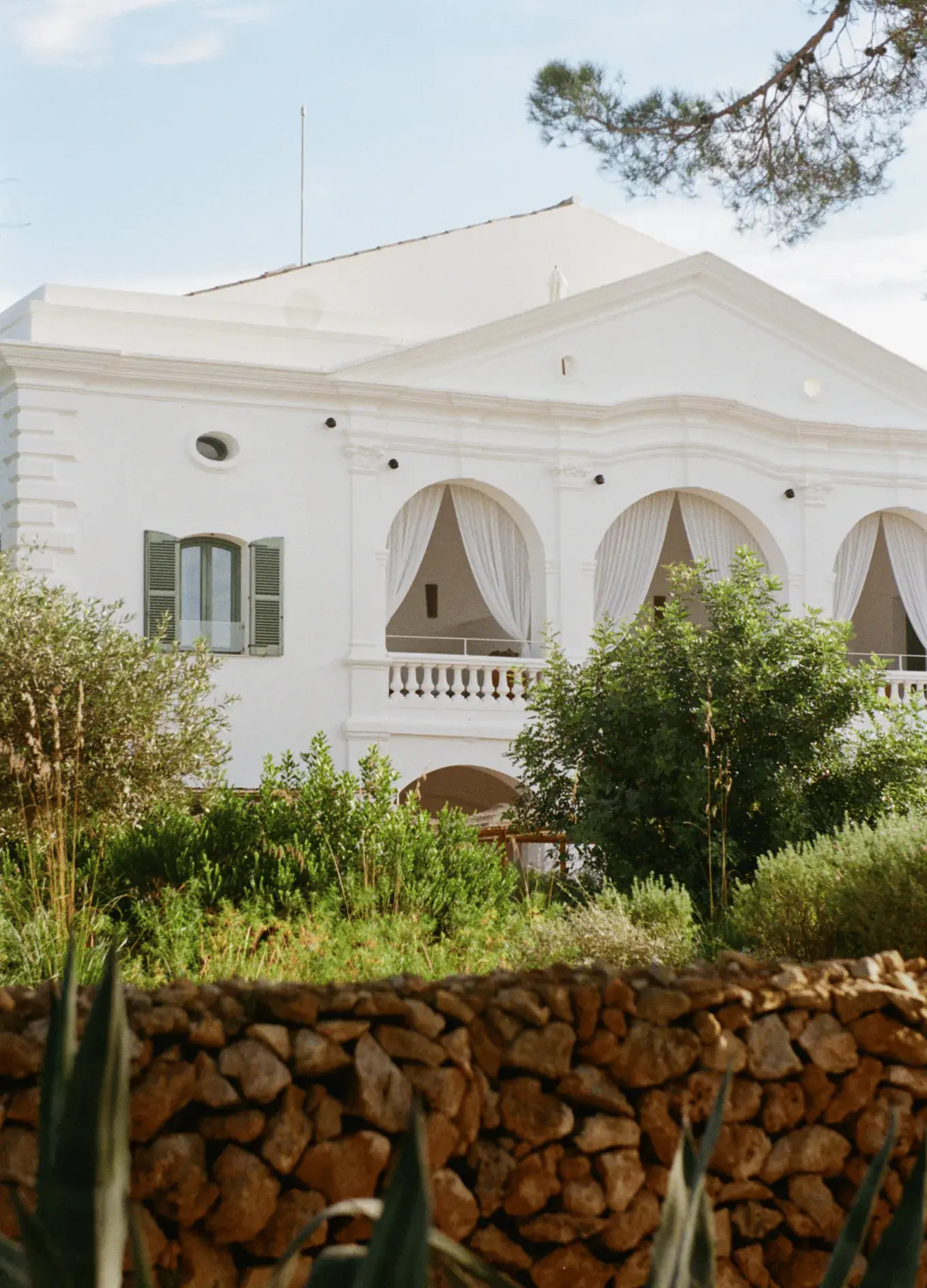 Edificio bianco in stile mediterraneo con balconi ad arco e persiane verdi dietro un muro di pietra e una vegetazione lussureggiante.