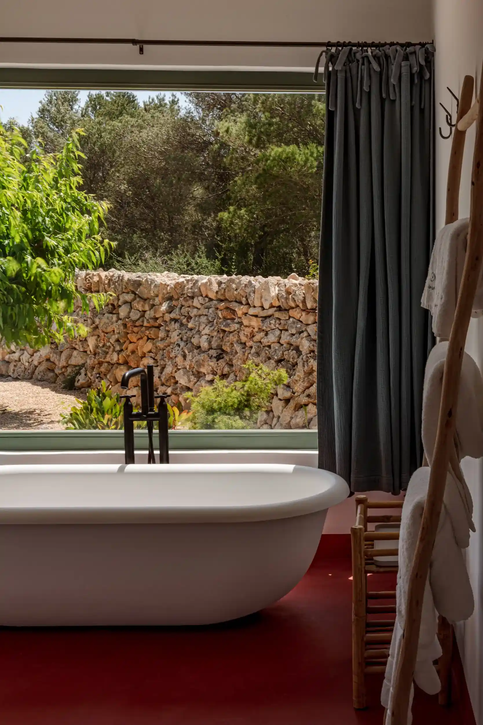 Vasca da bagno indipendente bianca con grande finestra che mostra pareti in pietra e vegetazione all'esterno in un bagno moderno.