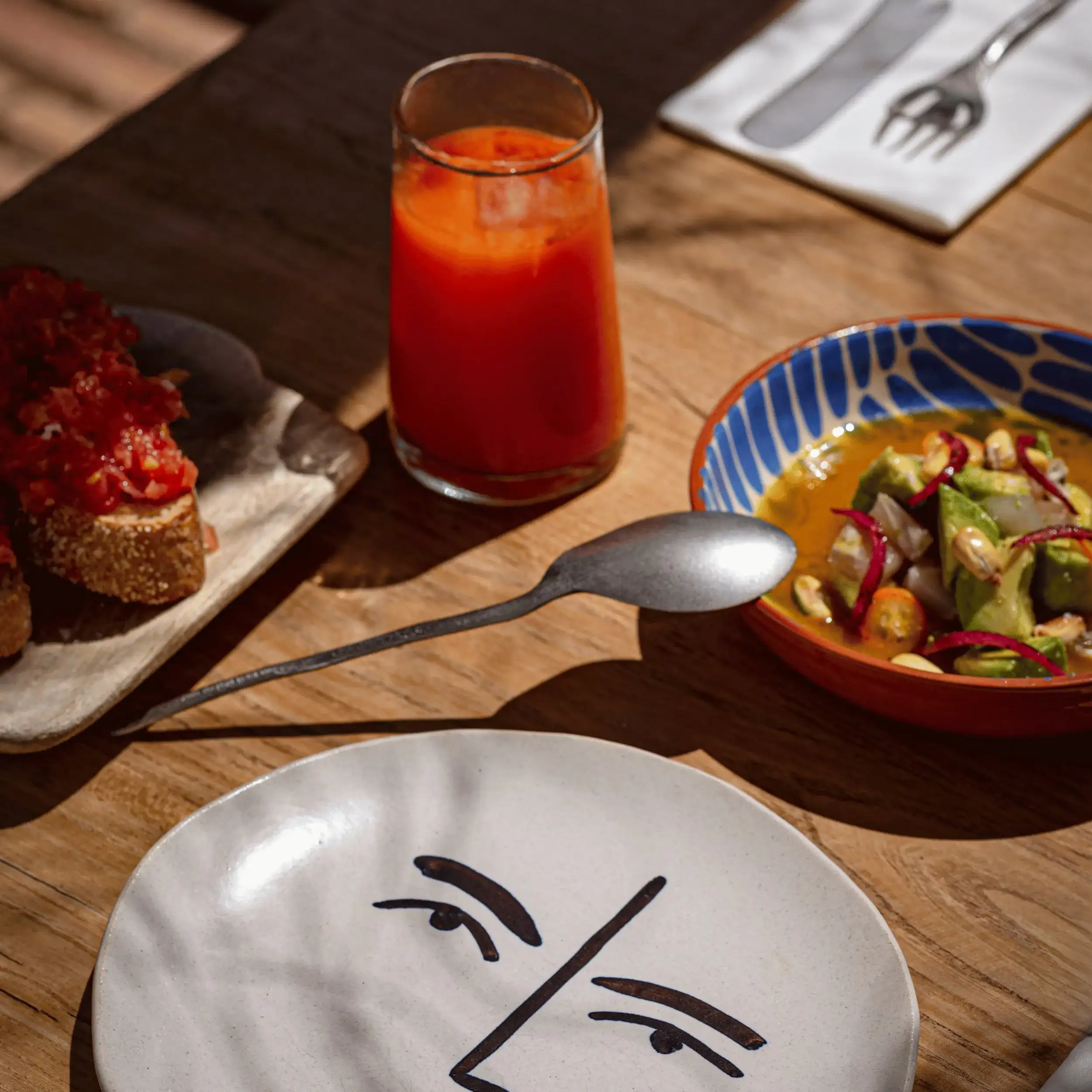Tavolo con un piatto a forma di faccia, un cucchiaio, un bicchiere di succo rosso, pane con pomodoro e un'insalatiera.