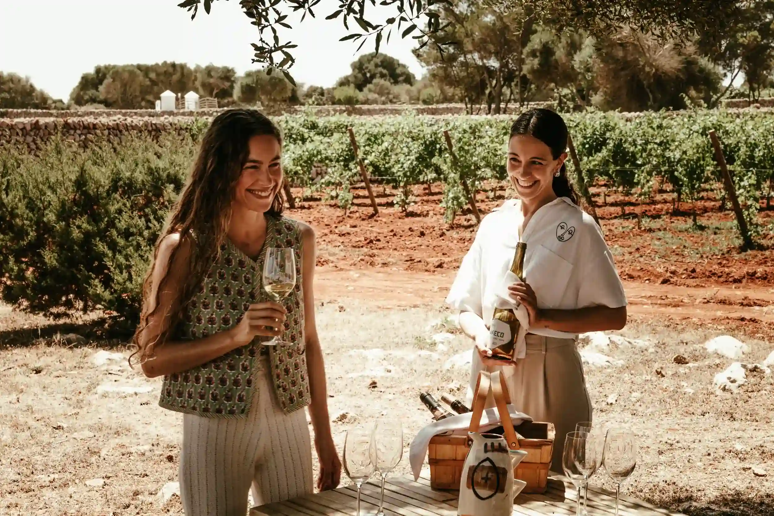 Due donne sorridenti all'aperto vicino al vigneto, una con in mano un bicchiere di vino, l'altra con una bottiglia di vino.