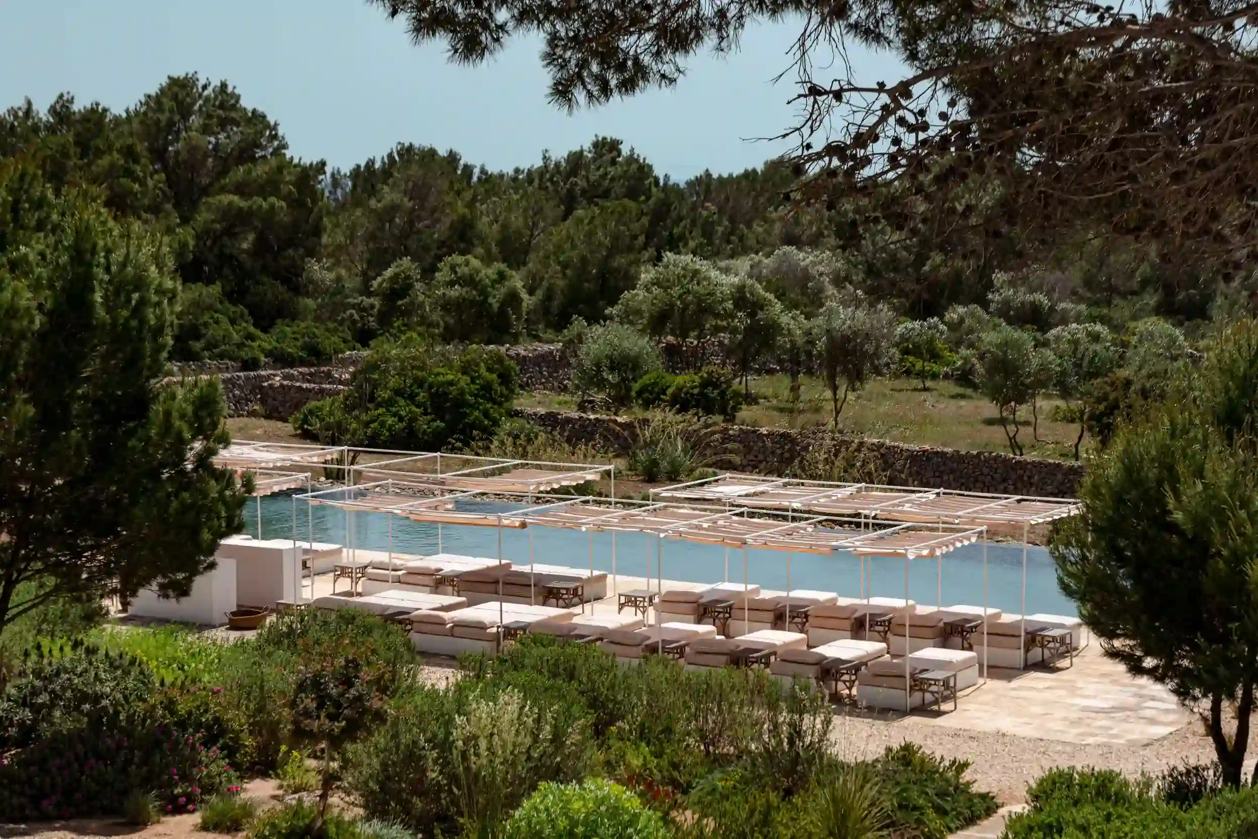 Zona piscina all'aperto con lettini imbottiti sotto pergolati bianchi circondati da alberi e arbusti.