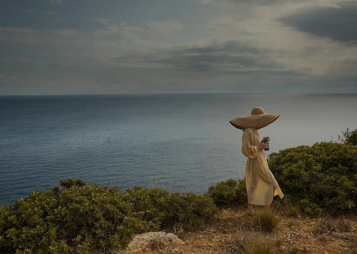 La persona in un abito beige e un grande cappello da sole si trova su una scogliera che si affaccia sull'oceano sotto un cielo nuvoloso.