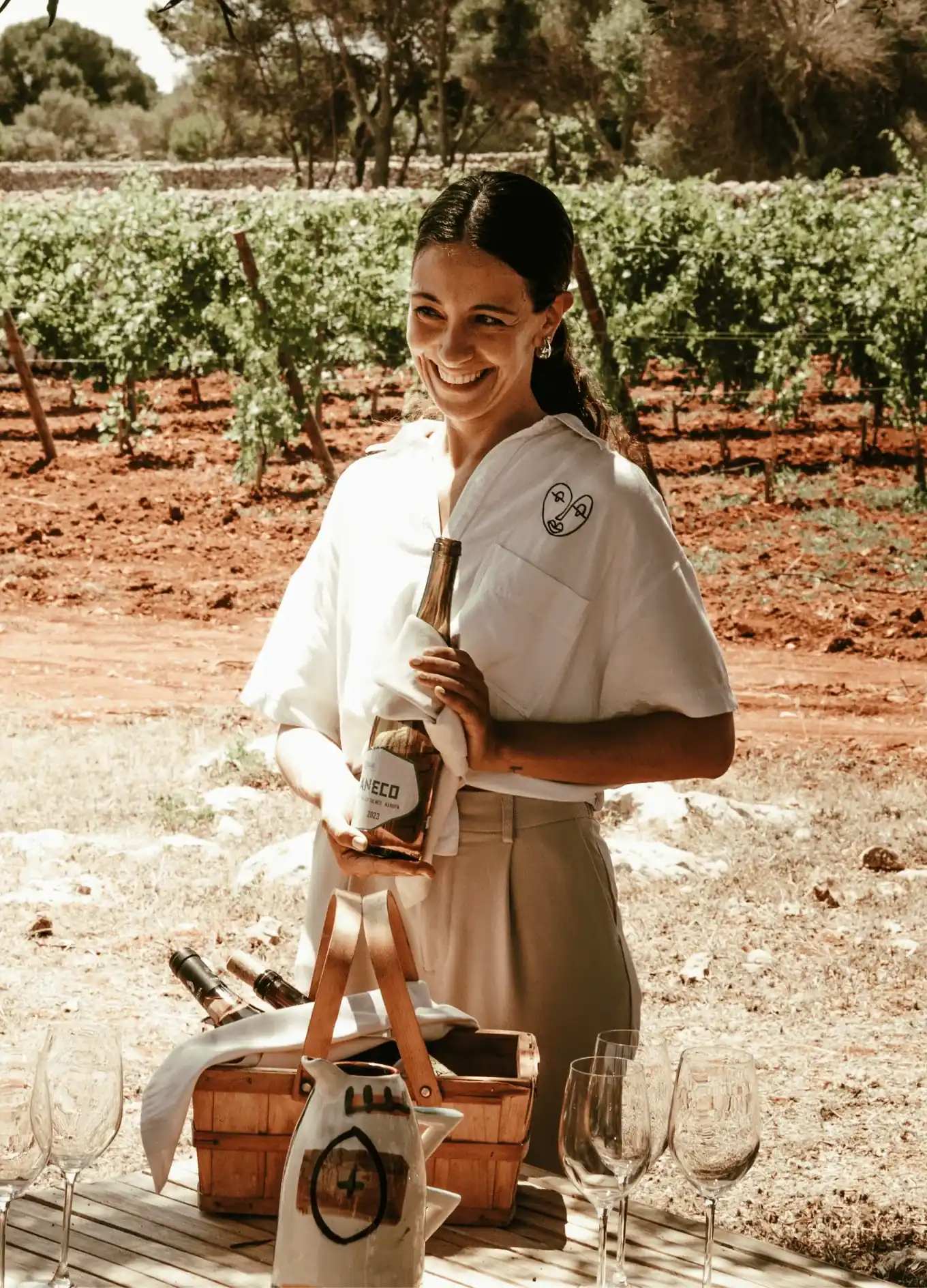 Donna sorridente che tiene una bottiglia di vino in un vigneto con bicchieri e un cesto di legno sul tavolo.