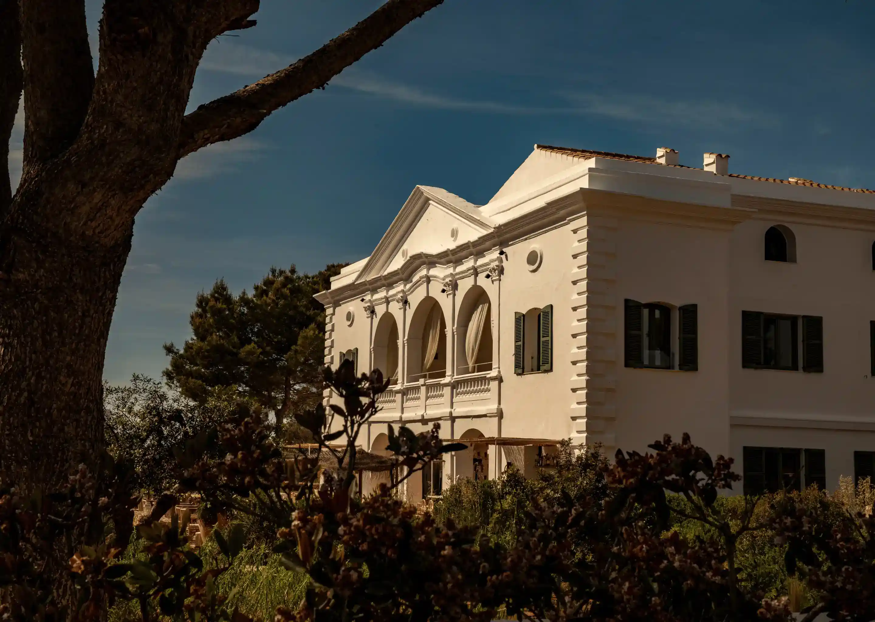 Edificio bianco classico con archi e persiane parzialmente oscurati da alberi e cespugli.