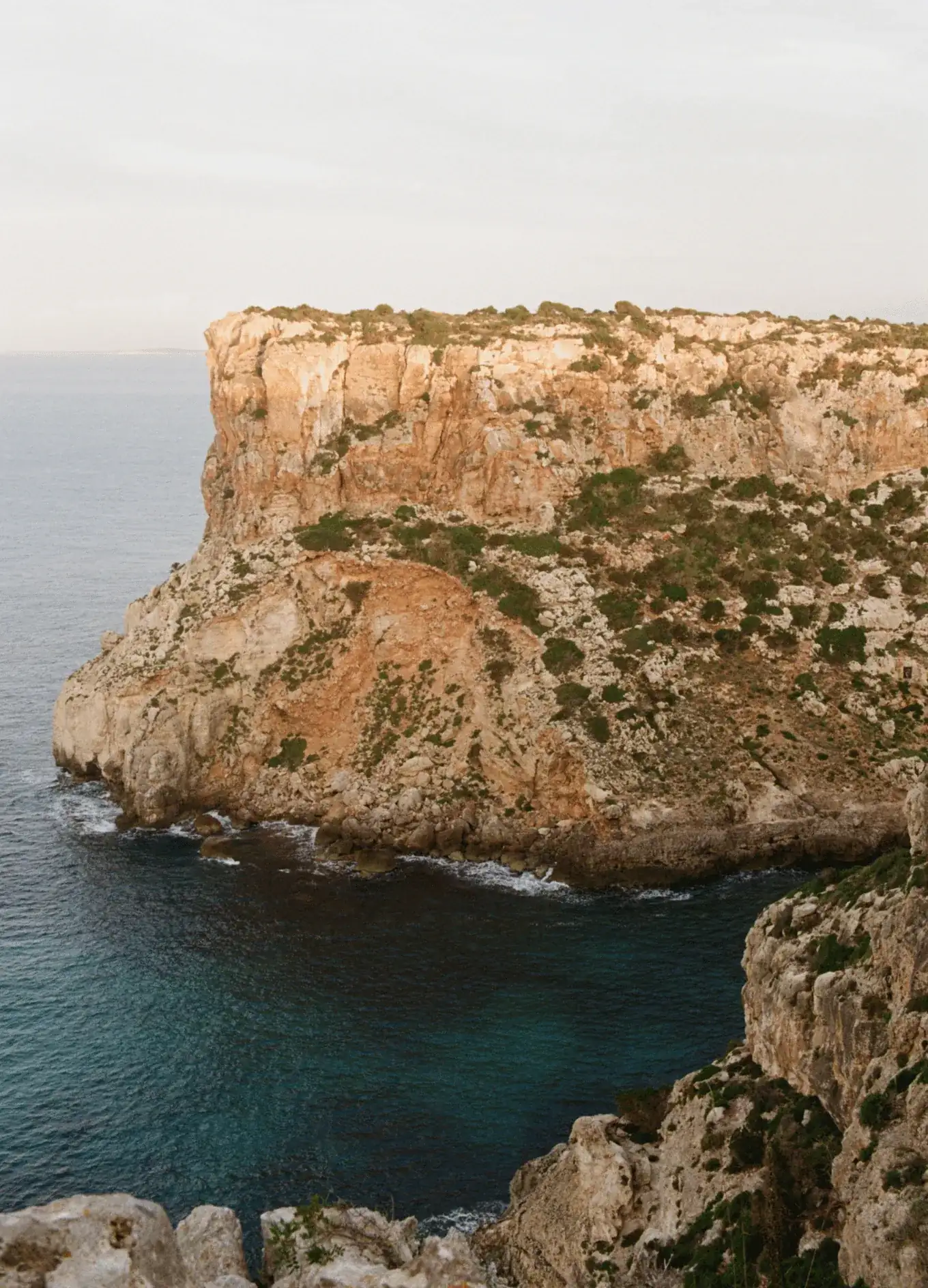 Scogliere rocciose ricoperte da una rada vegetazione in riva al mare blu intenso sotto un cielo nuvoloso.