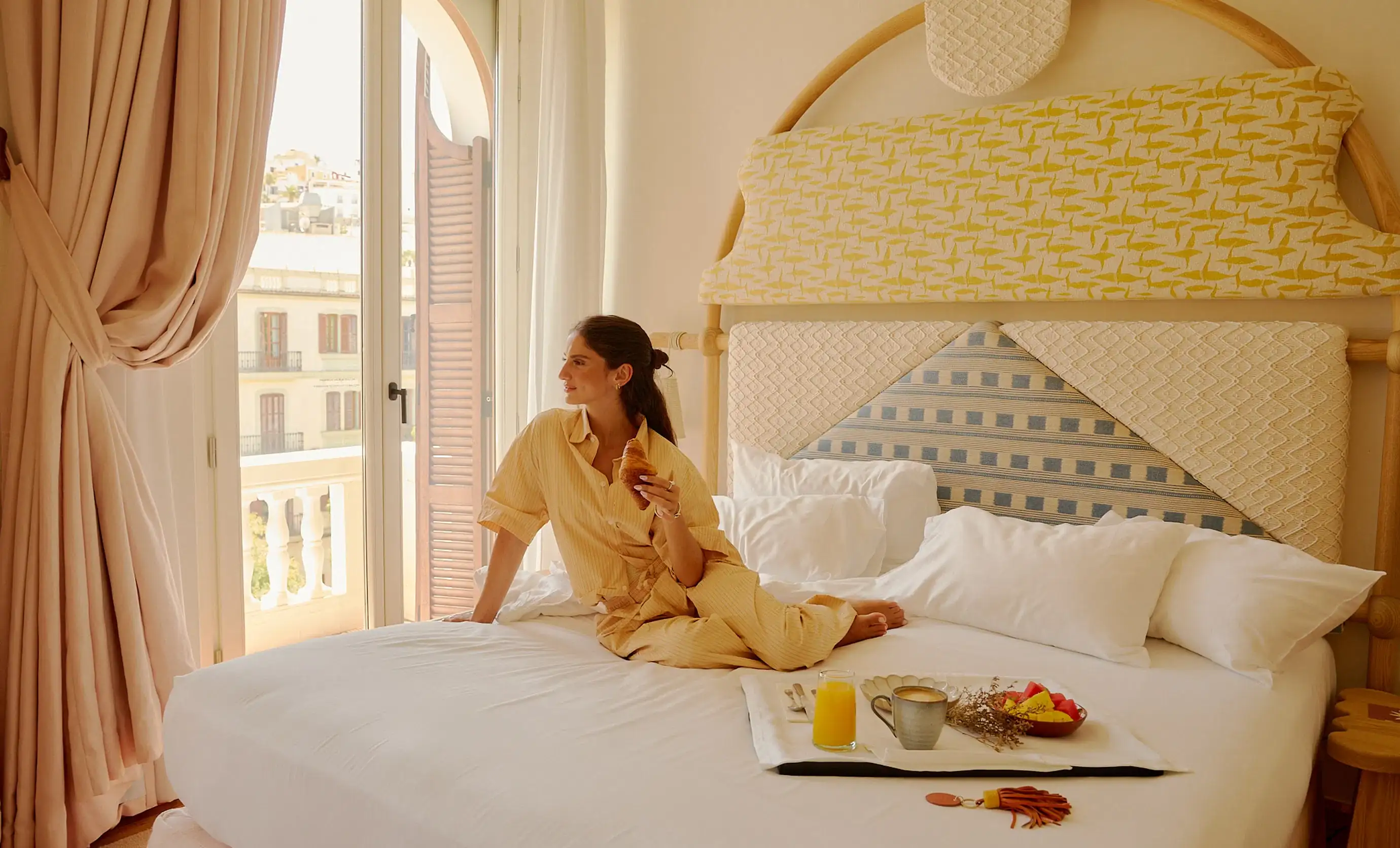 Mulher de pijama amarelo sentada em uma cama segurando um croissant perto de uma varanda com bandeja de café da manhã.