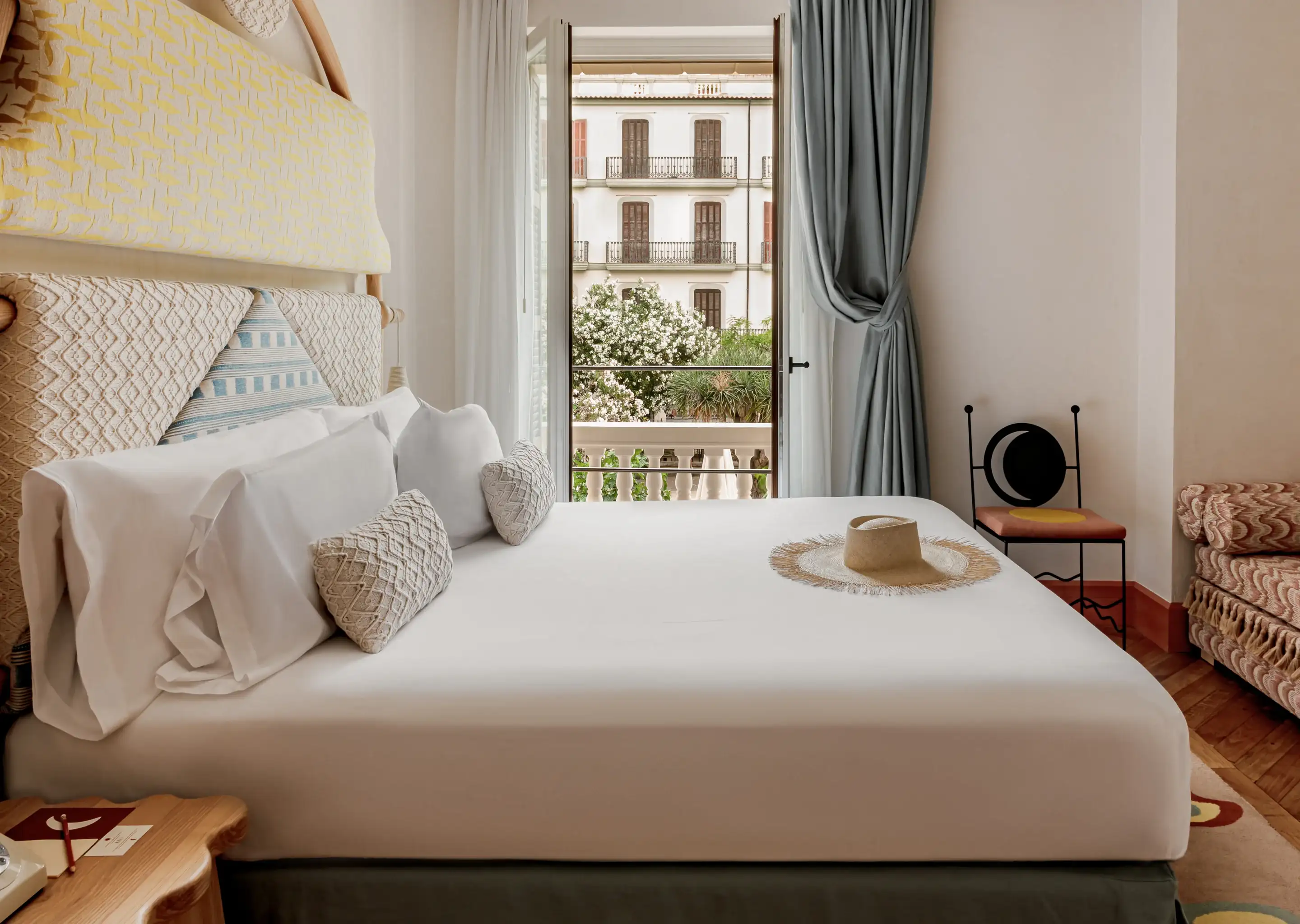 Quarto de hotel luminoso com uma cama grande, lençóis brancos, almofadas decorativas, chapéu de palha e vista para a varanda.