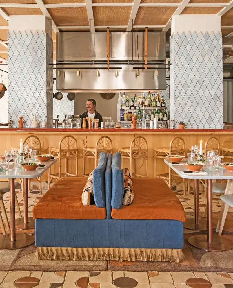 Interior de um bar com um barman sorrindo atrás do balcão, assentos e mesas de jantar com taças e copos.