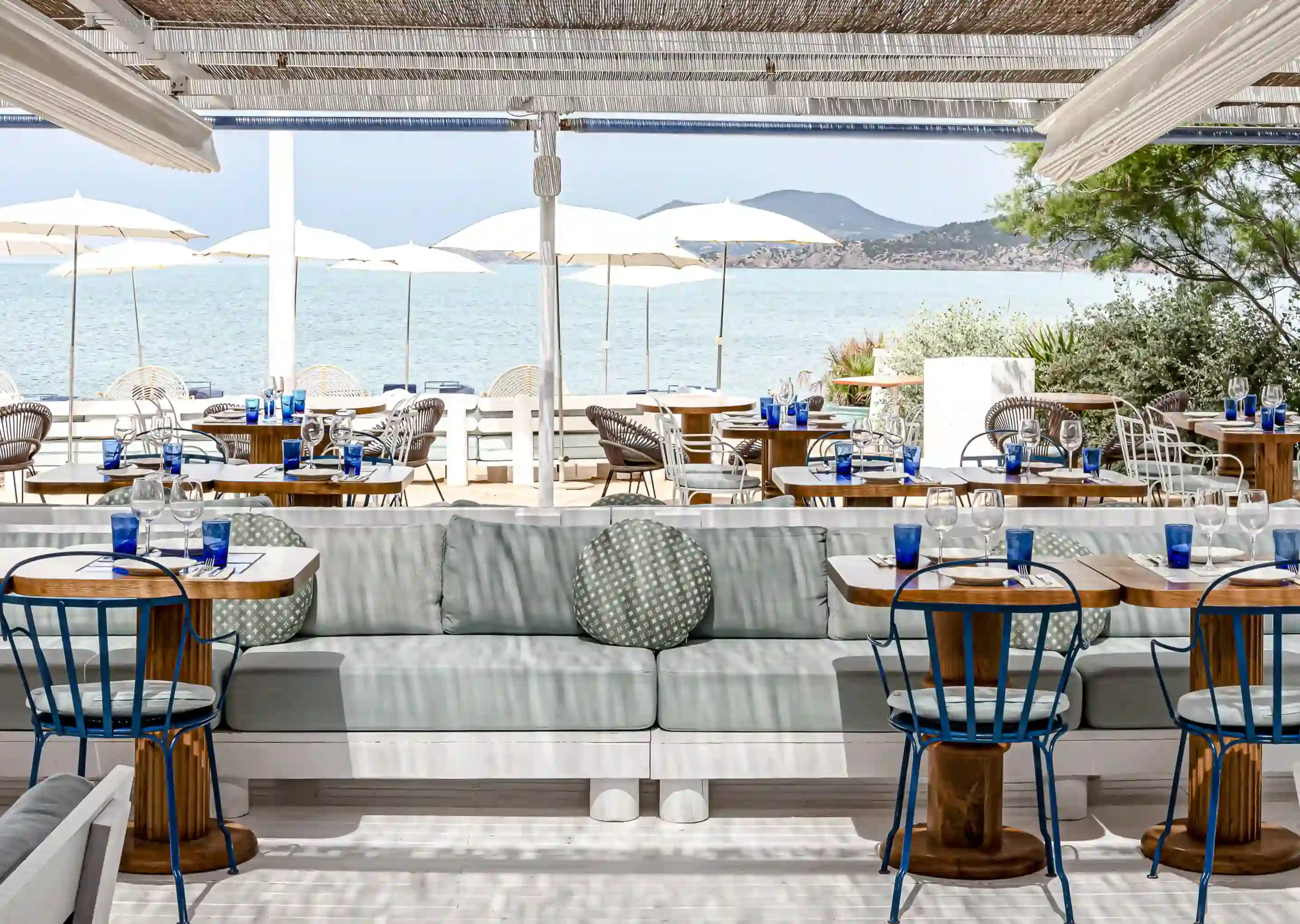 Restaurante ao ar livre à beira-mar com cadeiras azuis, mesas de madeira, guarda-sóis e vista para o mar ao fundo.