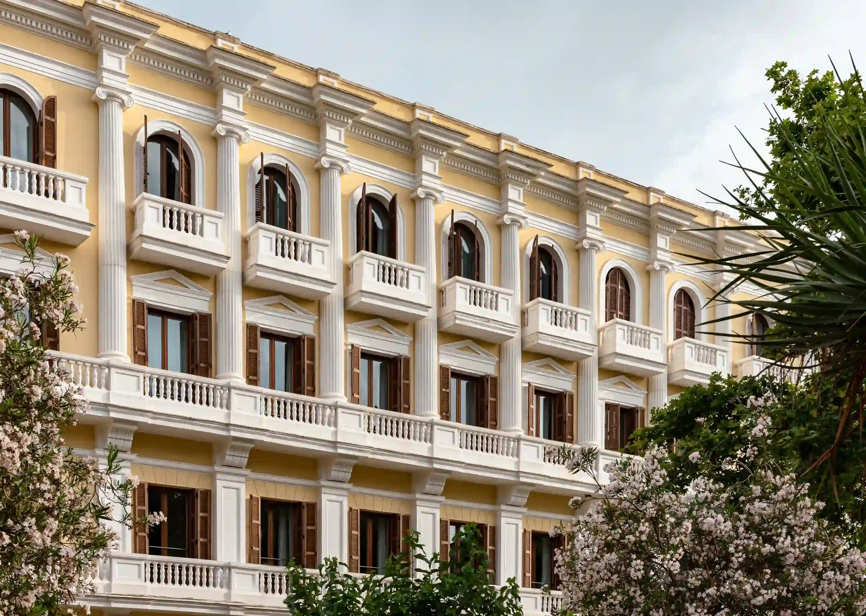 Fachada amarela do hotel com colunas e varandas brancas, cercada por árvores e flores verdes.