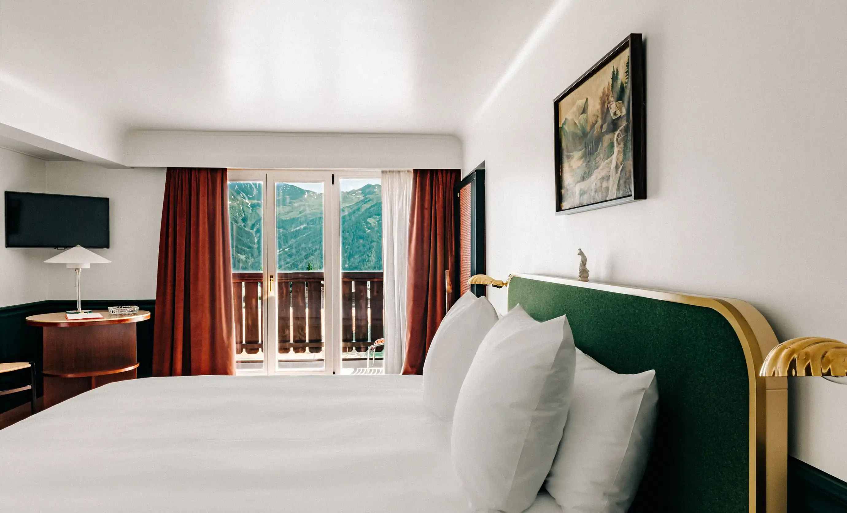 Quarto de hotel com cama branca, cabeceira verde, varanda com vista para a montanha, cortinas vermelhas e uma pintura de parede.