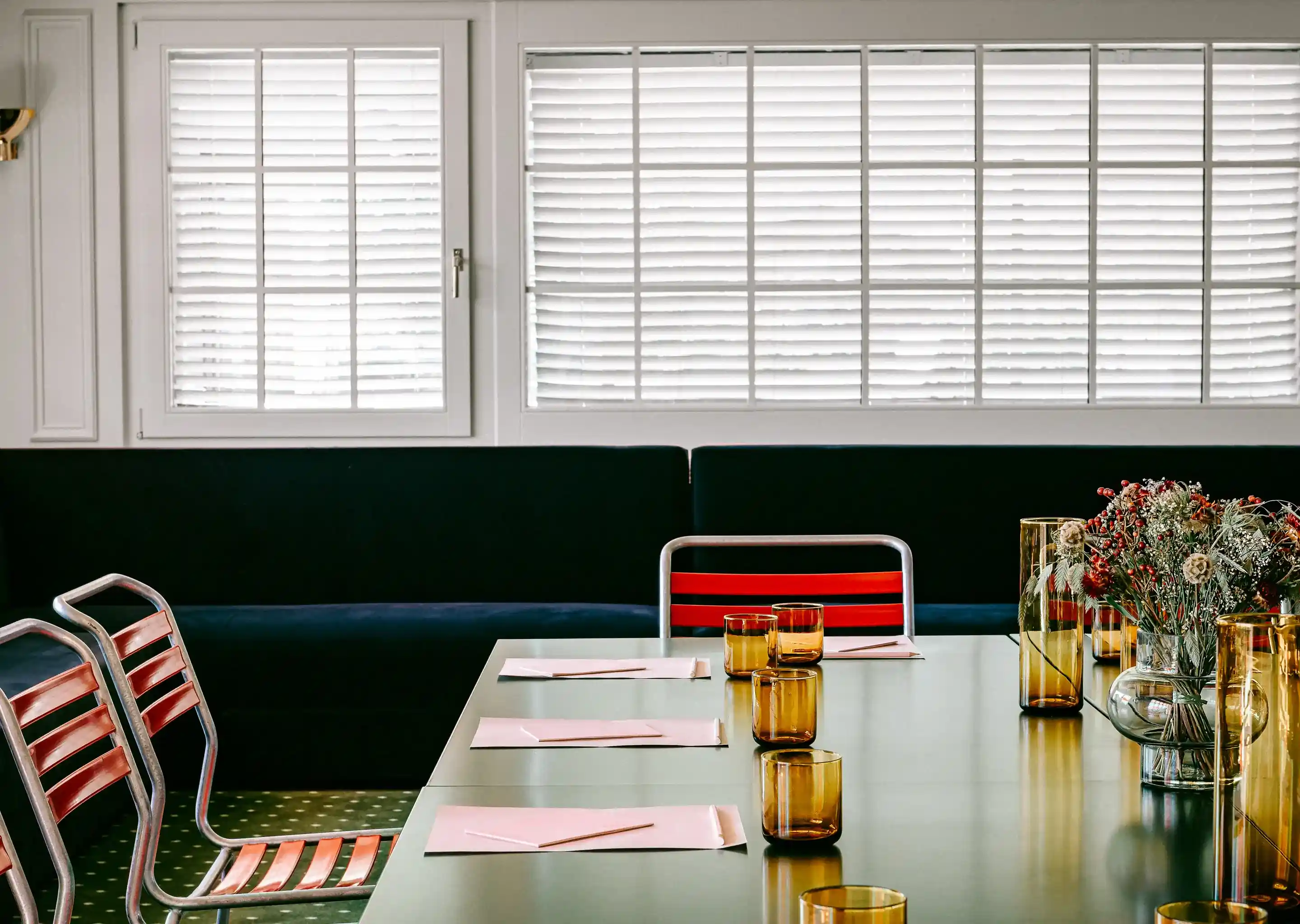 Conjunto de mesa para reuniões com copos de âmbar, papéis, lápis e peça central floral perto de persianas.