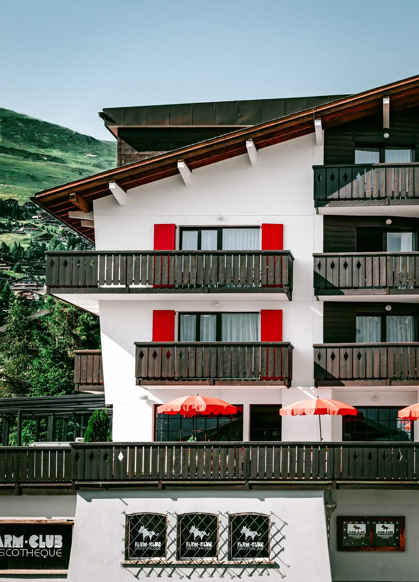 Edifício branco com varandas de madeira, persianas vermelhas, guarda-chuvas laranja e fundo verde na encosta.