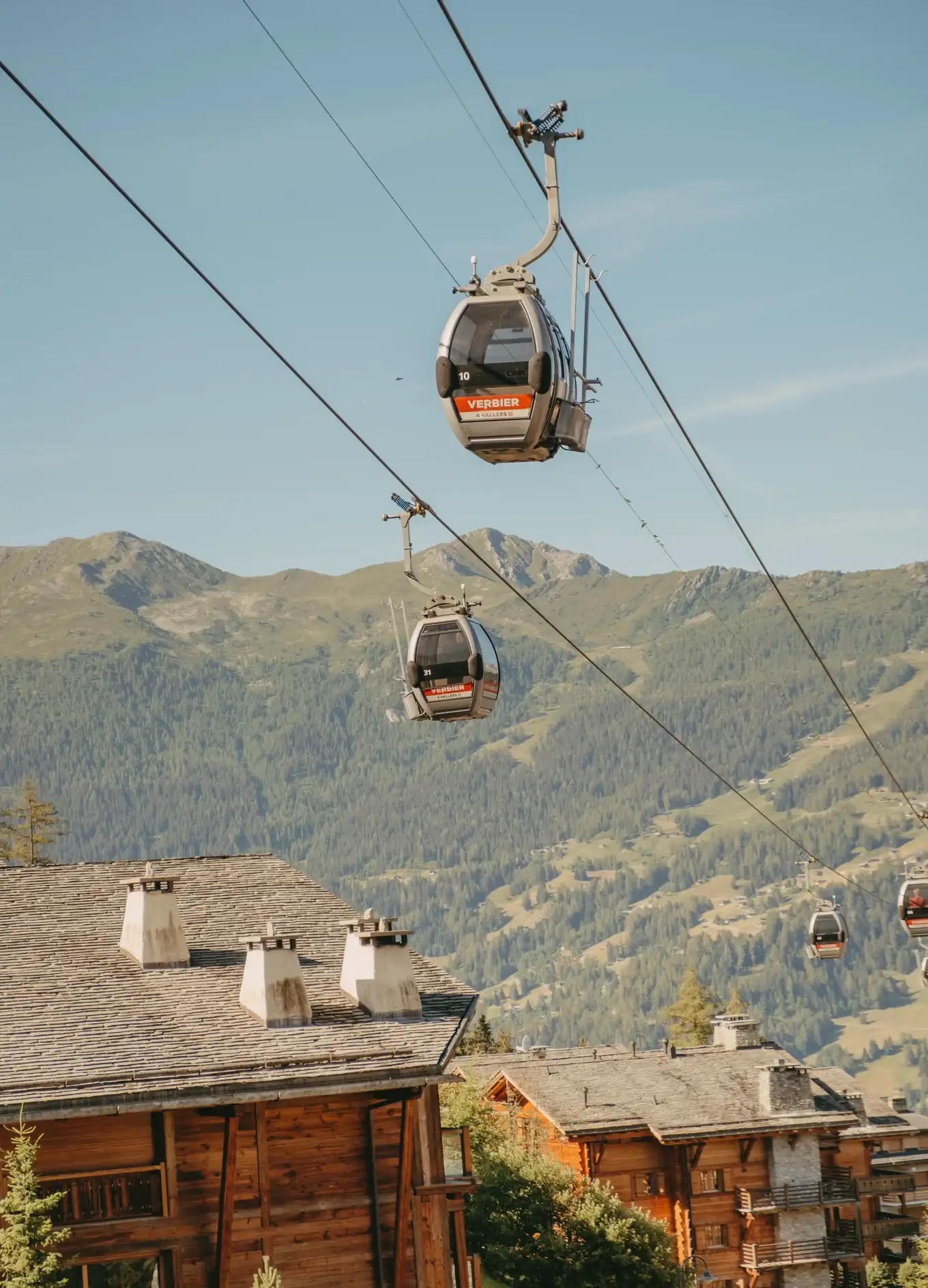 Teleféricos chamados Verbier se movendo acima de chalés de madeira com vista para a montanha ao fundo.