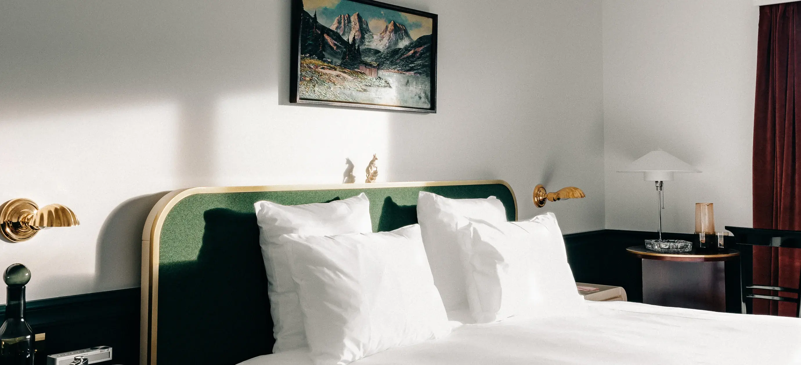 Cama de quarto de hotel com travesseiros brancos, cabeceira verde, luminárias de parede douradas, pintura e mesas laterais com luminária.