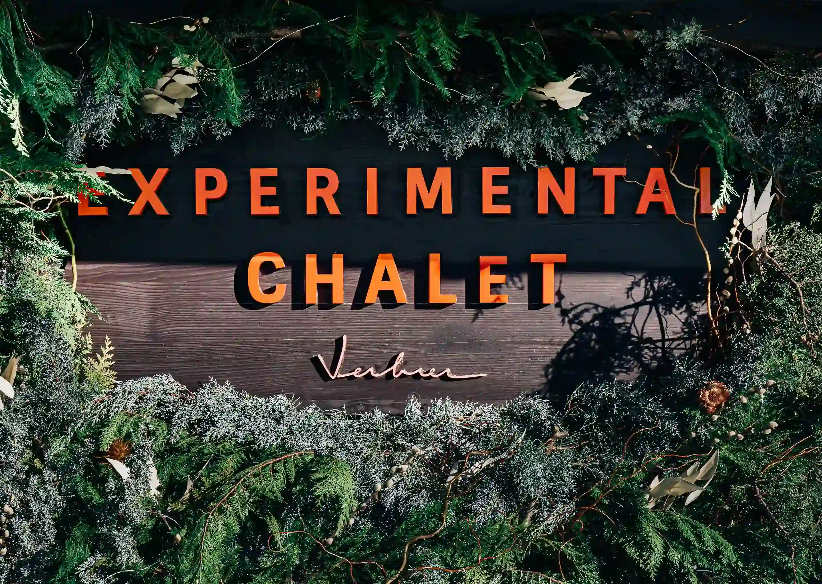 Placa onde se lê “CHALET EXPERIMENTAL Verbier” cercado por galhos e folhagens verdes de pinheiros.