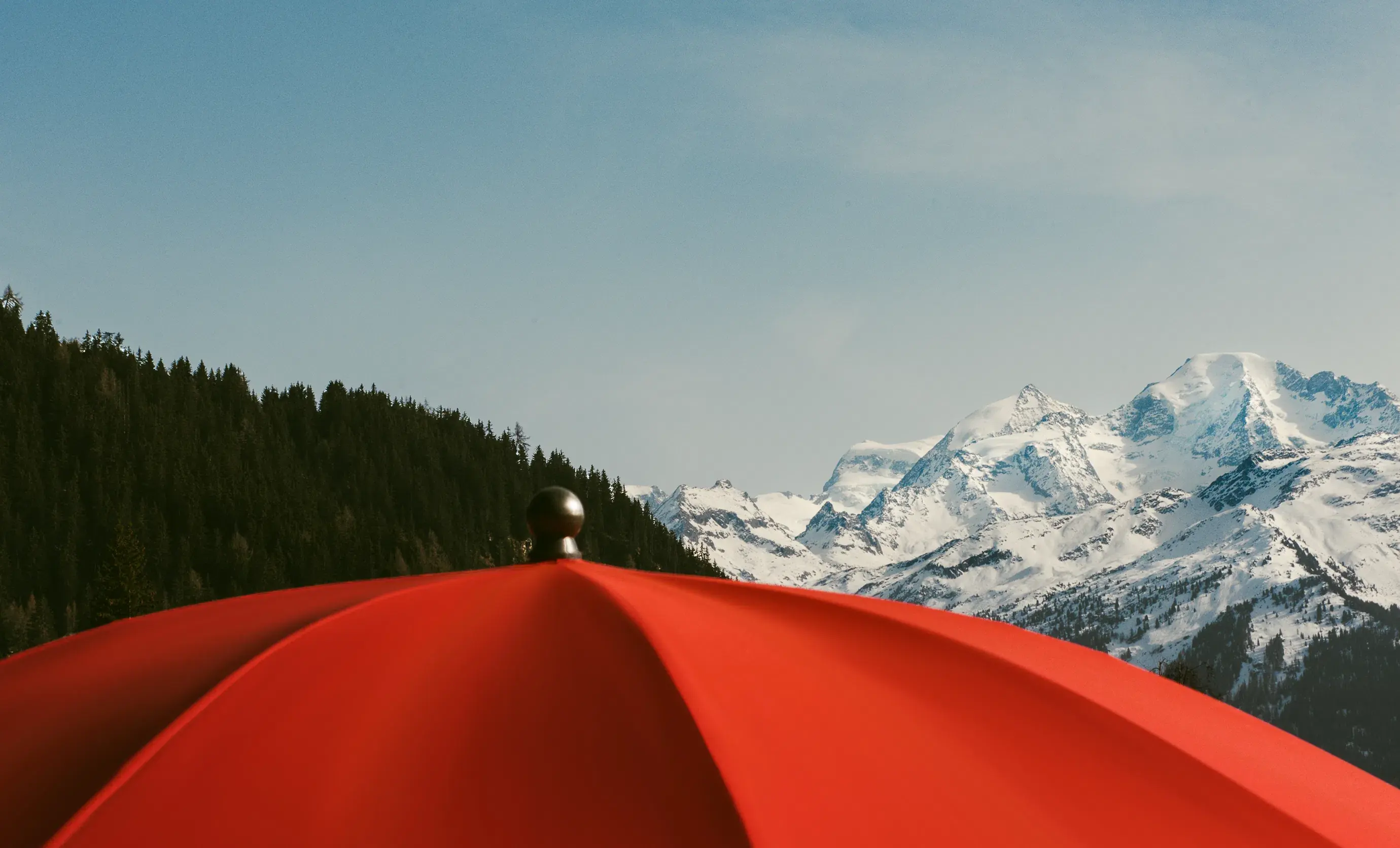 Guarda-chuva vermelho em primeiro plano com picos de montanhas nevadas e colinas arborizadas sob um céu azul claro.