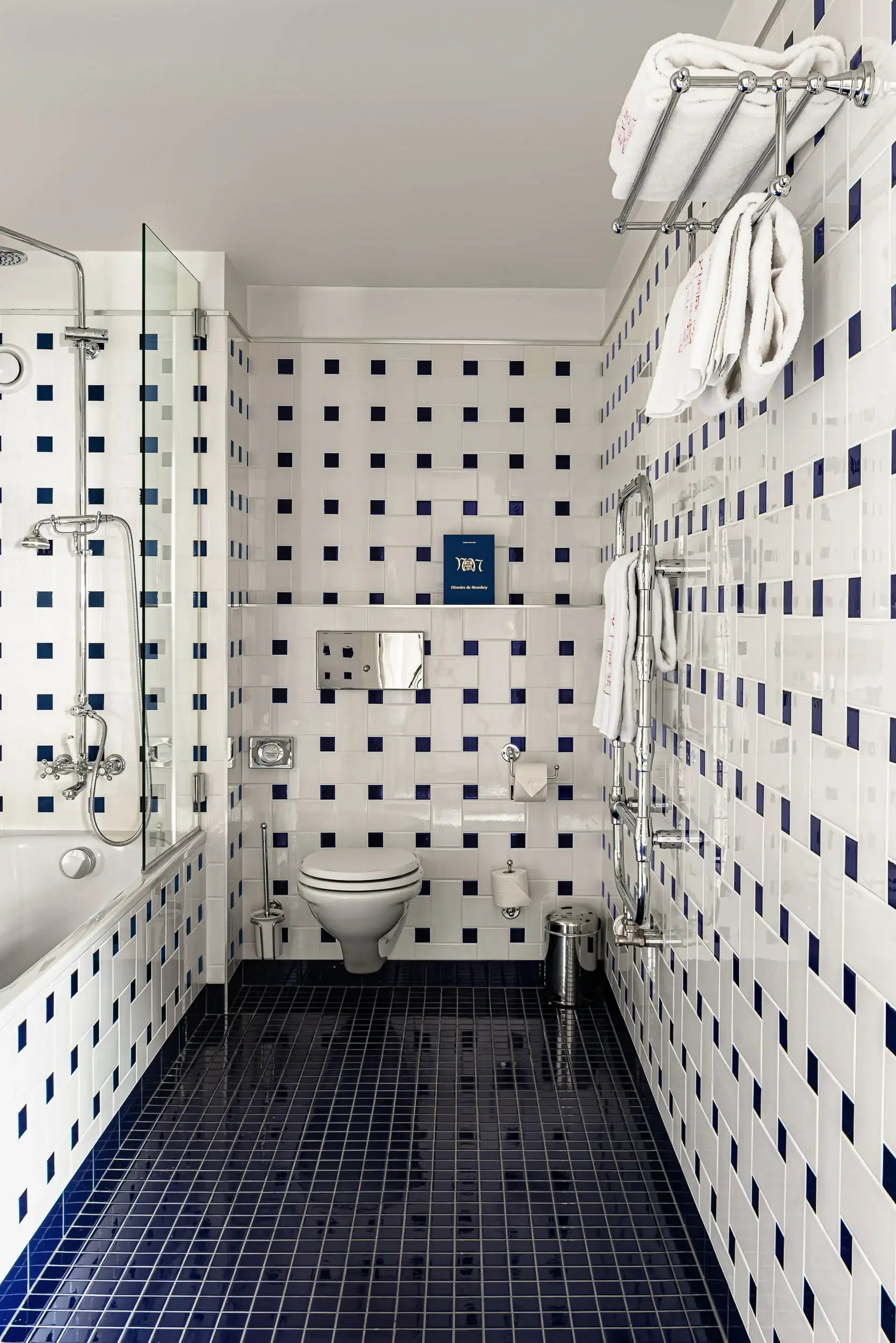 Banheiro com paredes e piso de azulejos brancos e azuis escuros, banheira, vaso sanitário e toalheiro.