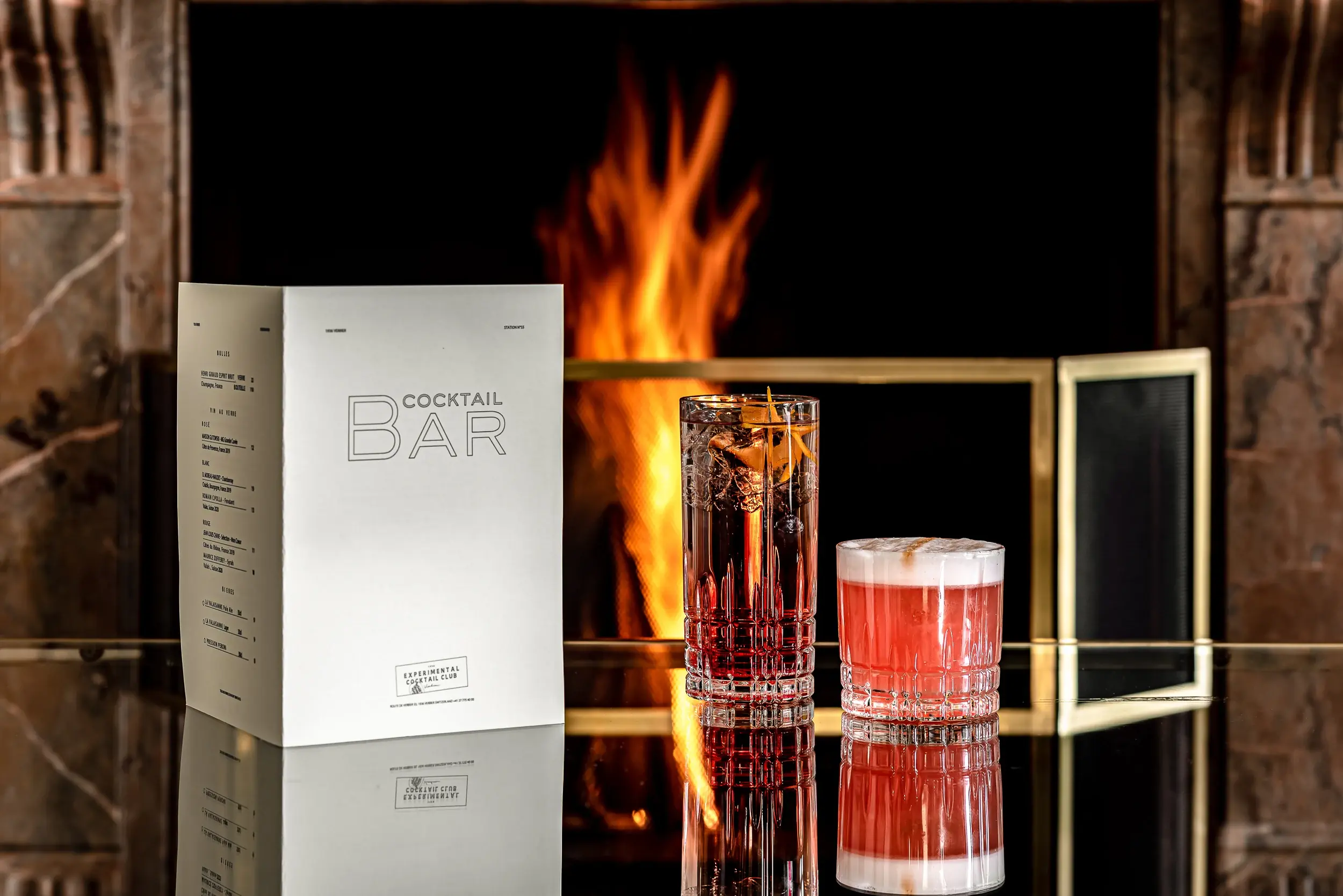 Dois coquetéis e um menu de bar de coquetéis em uma superfície refletiva em frente a uma lareira aconchegante.